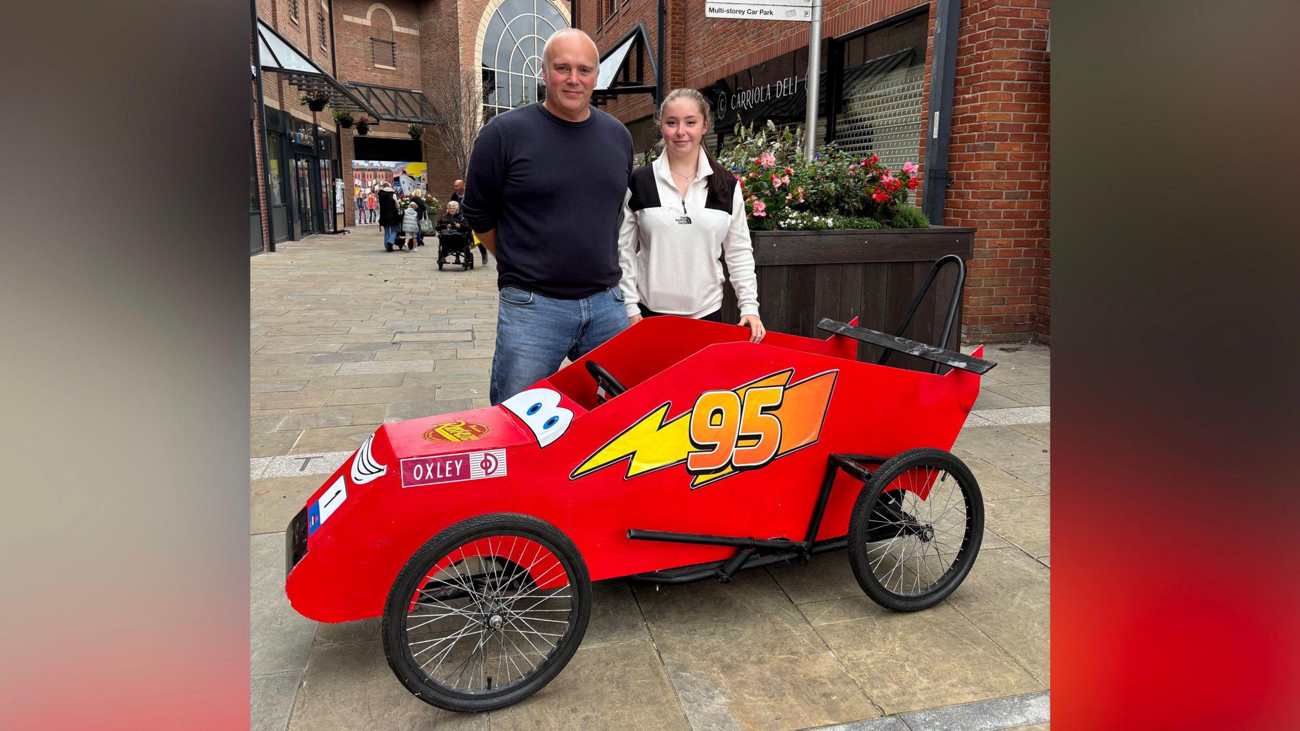 Two people stand smiling in front of a red cart, made up to look like the Cars character Lightning McQueen.