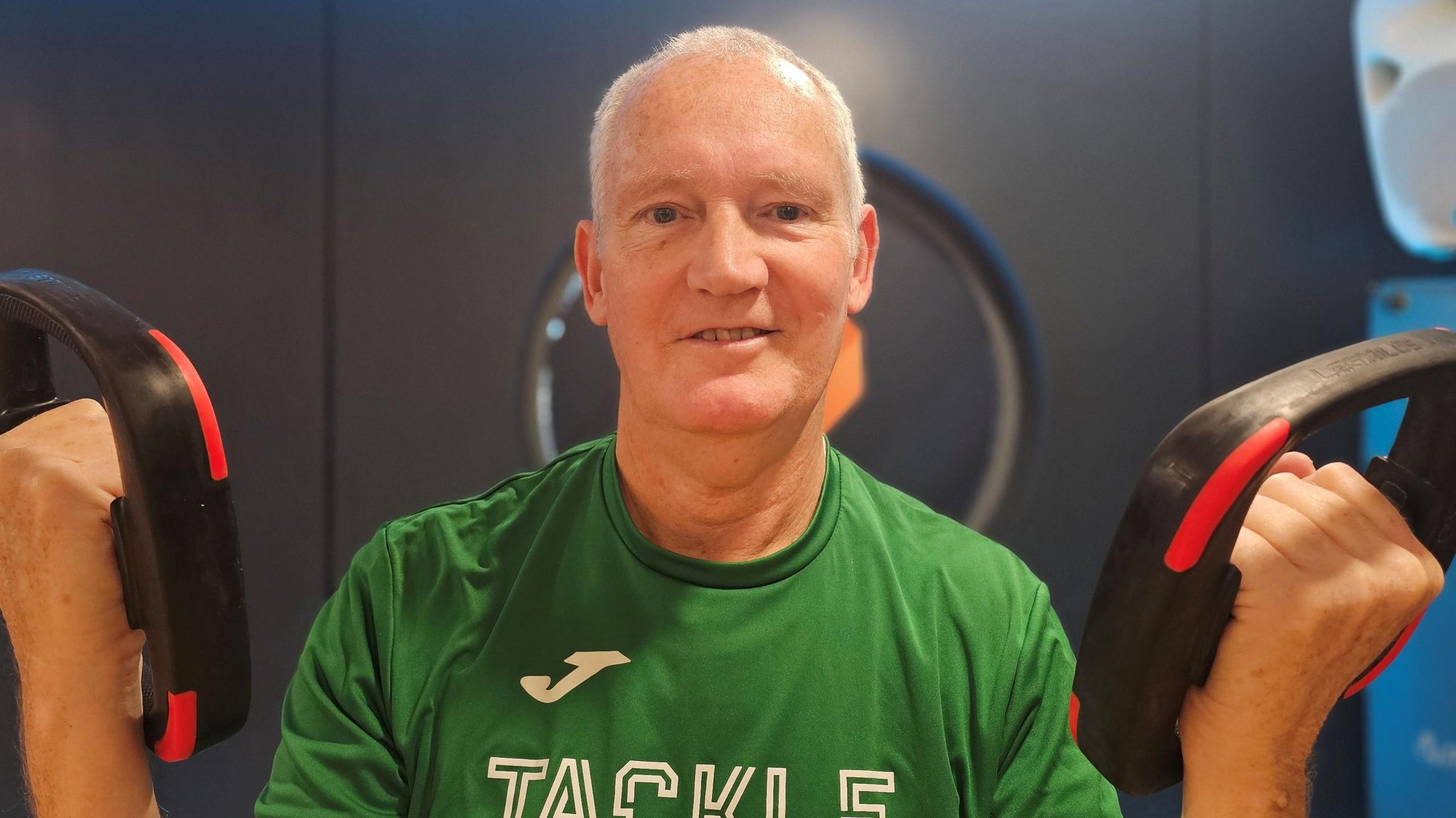 Steve Stone - who has short white hair - wearing a green t-shirt with the word 'Tackle' on the front in white capital letters. He is holding a piece of workout equipment in each hand.