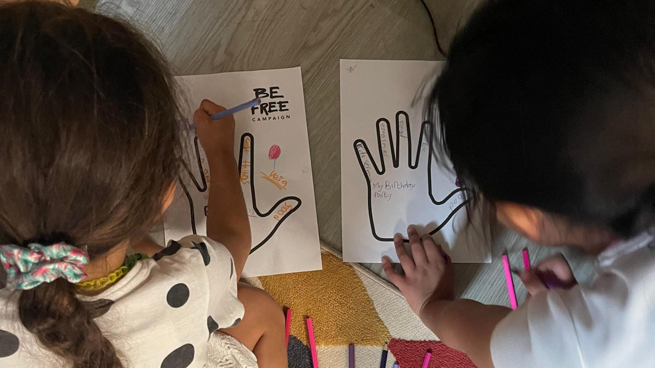 A picture of two young children completing a "memory hand" exercise. The picture is taken over their shoulder and shows them writing positive memories inside the outline of a hand.
