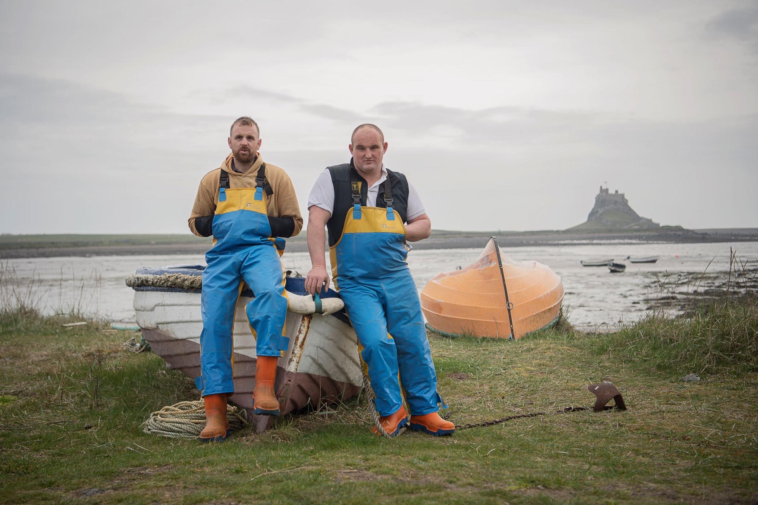 Two fishermen in blue overalls lean either side of the bow of a small fishing boat, grounded on grass beside the sea, with a small castle across the water in the distance.