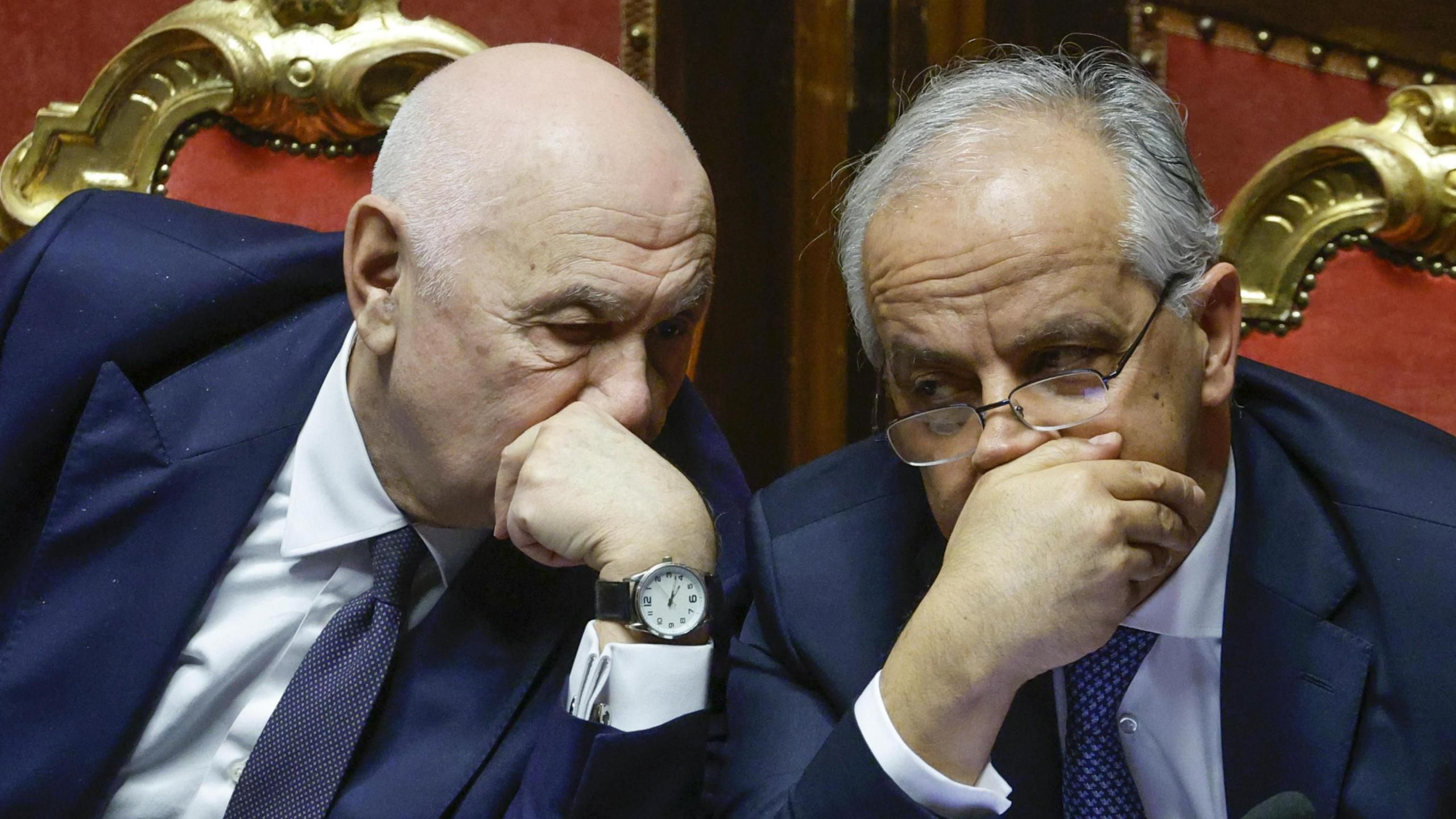 Carlo Nordio, wearing a watch and a dark tie, talks to his ministerial colleague who is wearing glassses, while both talk behind their hands.