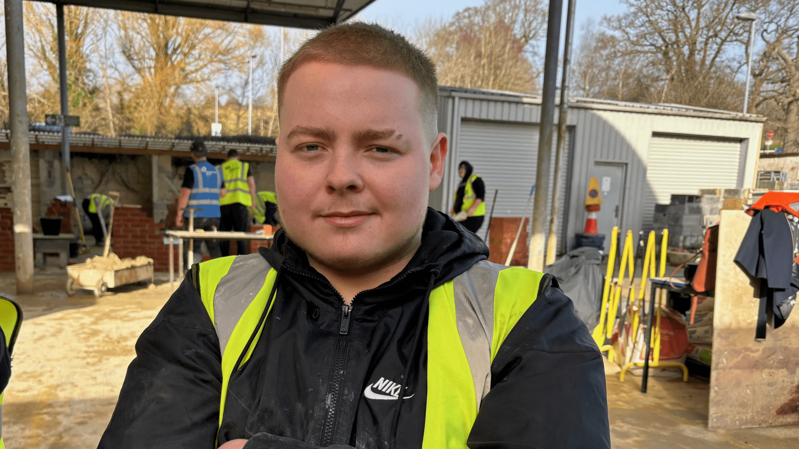 Head and shoulders shot of Will West looking at the camera in a yellow high-viz jacket over a black jacket, sat in a brick yard. 