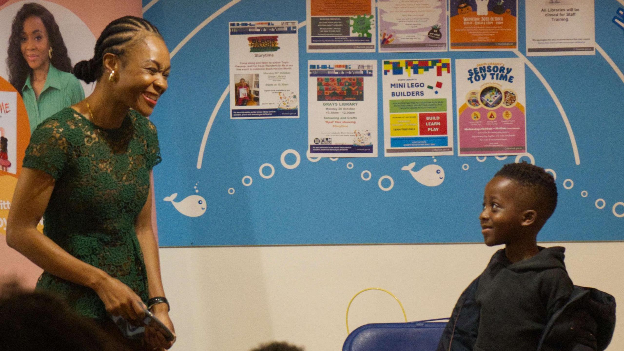 A woman wearing a green dress smiling at a boy who is smiling back, looking slightly shy. He is wearing a black hoodie while his coat hangs off him. They are both standing in front of an information board at a library.