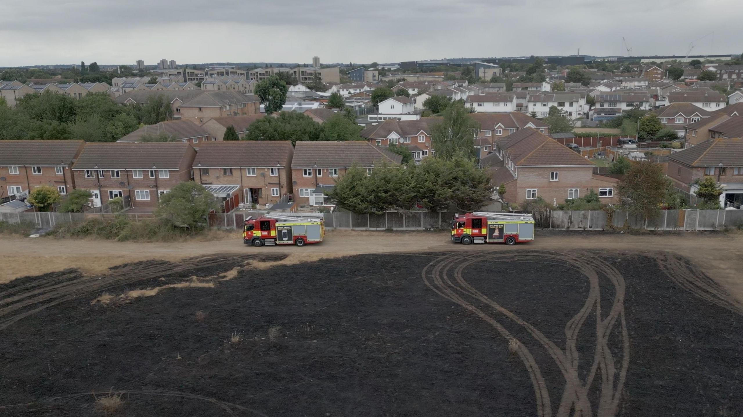 Two fire engines are parked on the edge of a burnt out field adjacent to a row of houses. They must have driven on the field as you can see track marks on the brown, darkened ground. 