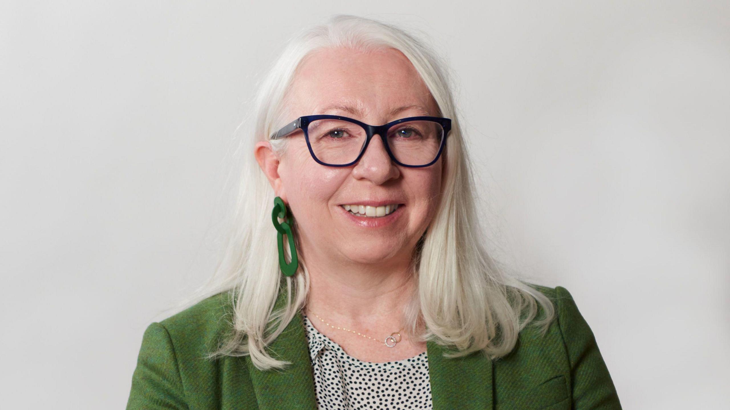 Anne Lyden looking directly at the camera. She has white, shoulder length hair and is wearing glasses with a dark frame. She has long, green earrings on. She is wearing a green blazer and a white and green dotted top. She is standing in front of a neutral background.