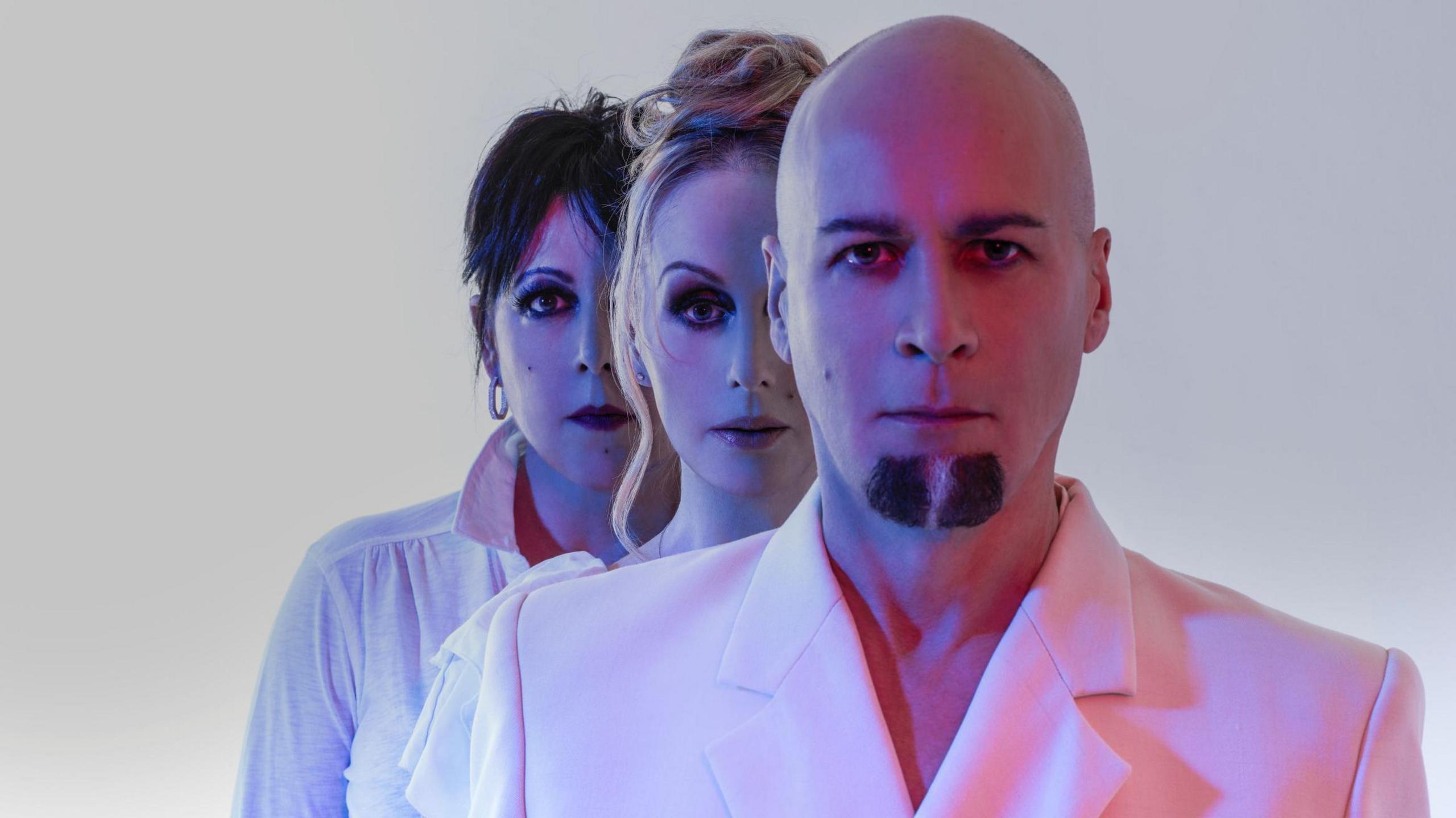 The three band members stand in a row in pink and purple lighting