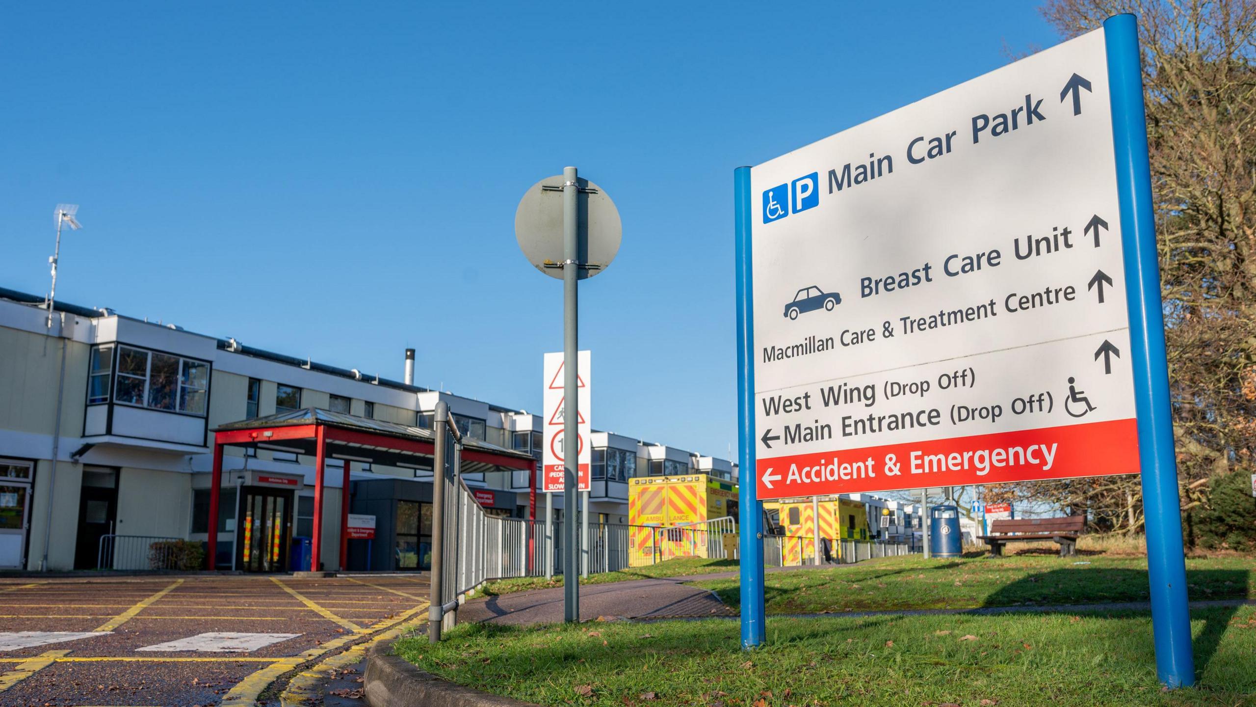 Outside the Queen Elizabeth Hospital in King's Lynn. A sign can be seen that is signposting people to various places, including the main car park, breast care unit, Macmillan Care and Treatment Centre, the West Wing, main entrance, and accident and emergency departments. Two yellow ambulances can also be seen.