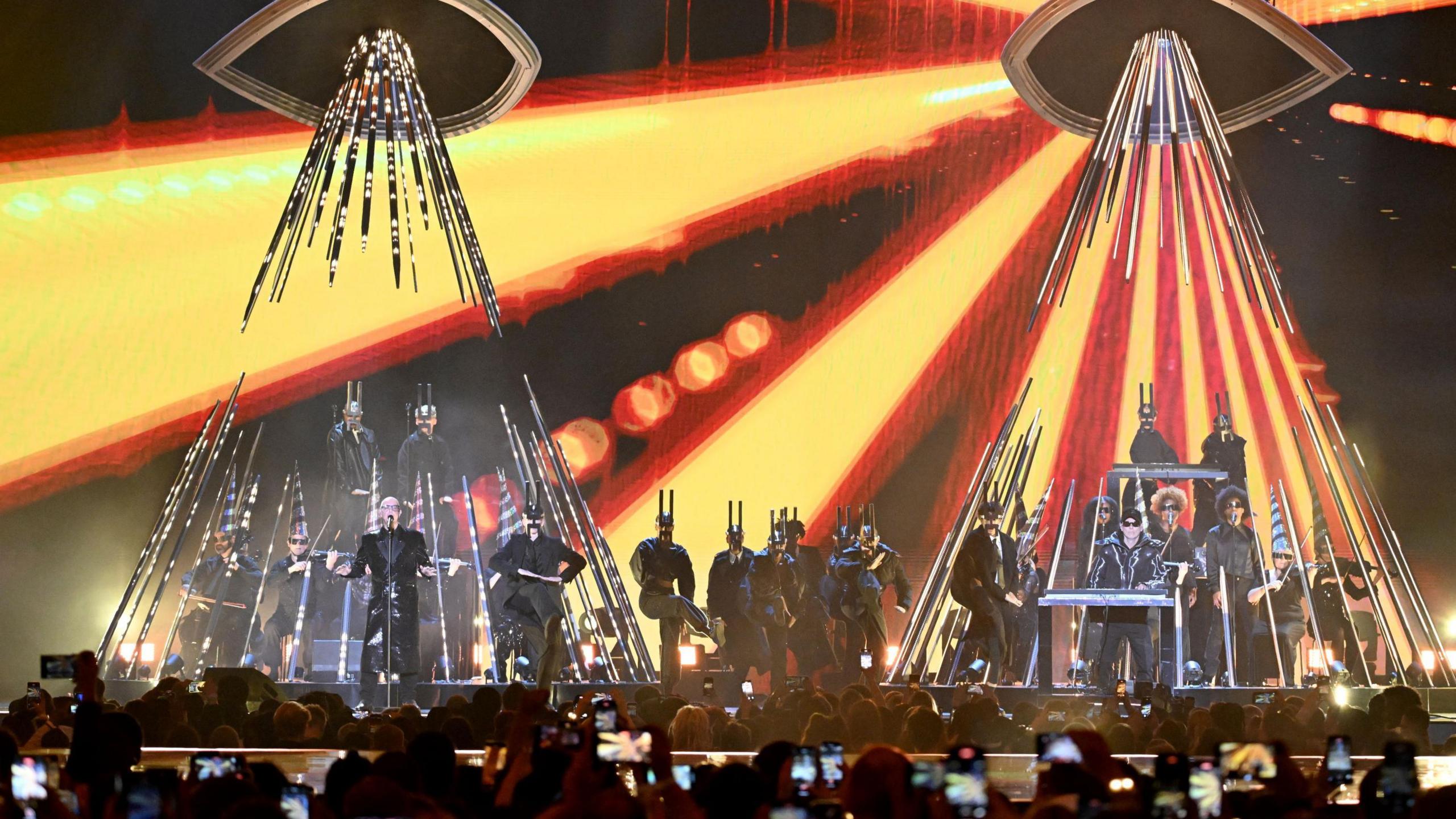 The Pet Shop Boys performing a show, with bright lights shining down on them and musicians in silver costumes behind.