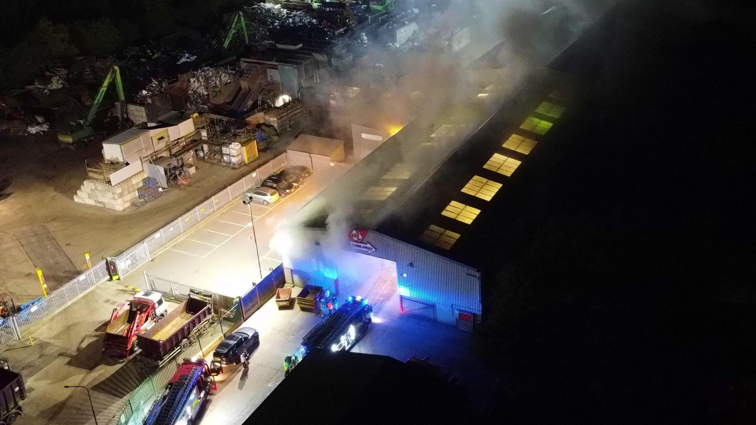 Smokes pours out the windows and doors of a warehouse at night. 