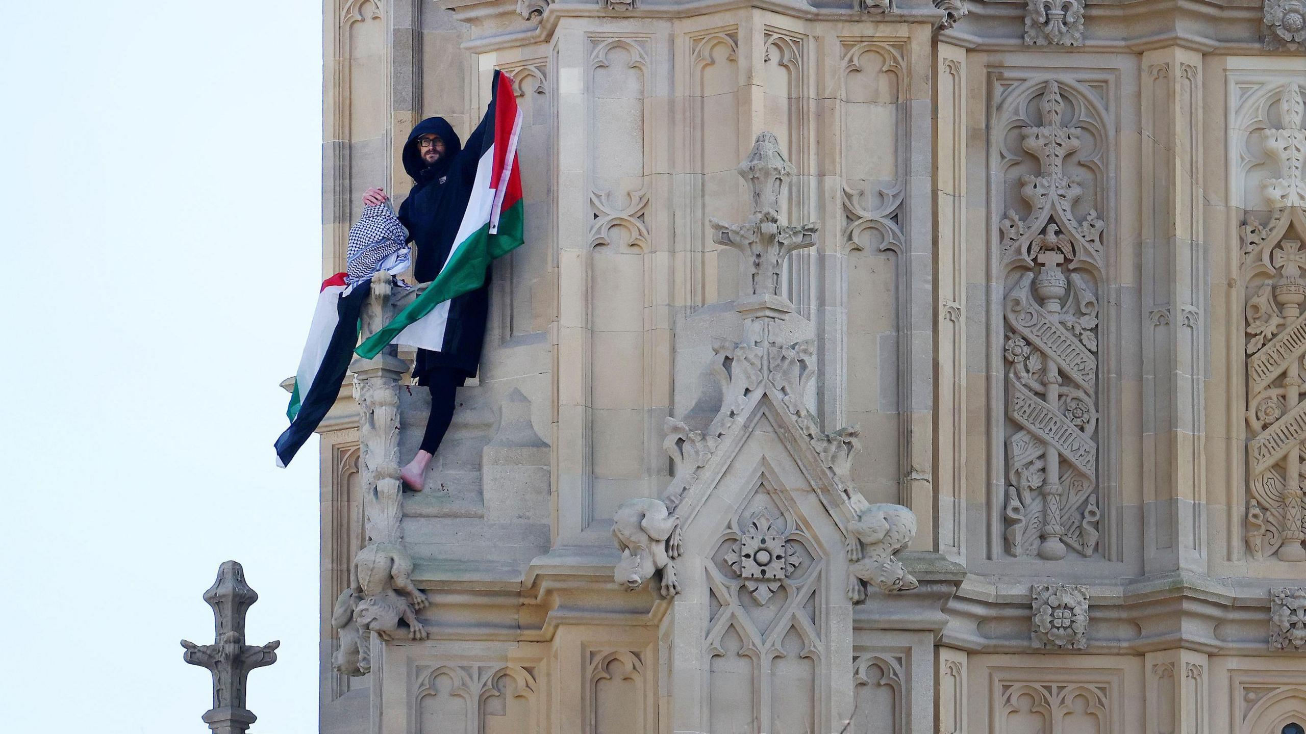 A pro-Palestinian protester waves a Palestinian flag atop the Elizabeth Tower. The man, dressed in black hoodie and joggers is resting against a ledge and holds the flag aloft with his left arm. He is barefooted with his left foot resting on a stone gargoyle.