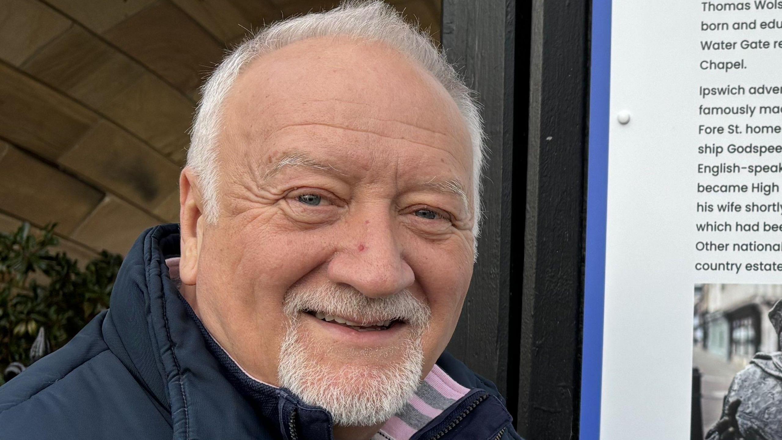 A man with grey hair is smiling at the camera. He is wearing a navy gilet jacket with a navy jumper underneath and a white top. He is standing in front of a town information board detailing its history and points of interest.