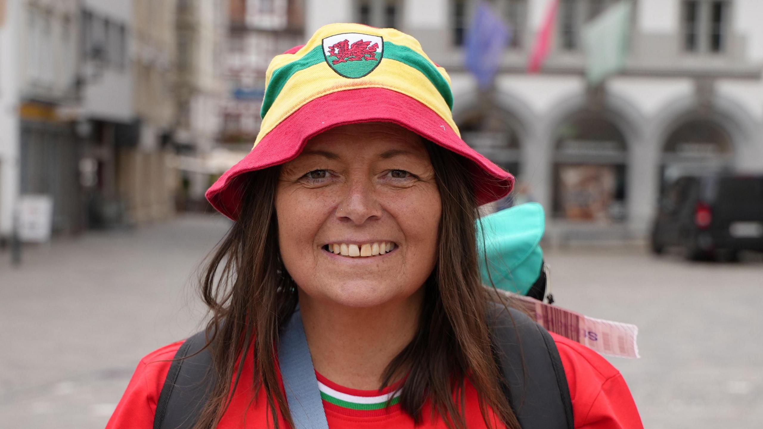 Lisa Folkes wearing a red Wales shirt and smiling at the camera. She is wearing a red, yellow and green bucket hat with the Welsh dragon on the front, and carrying a green bag. She is standing in front of a building with flags.