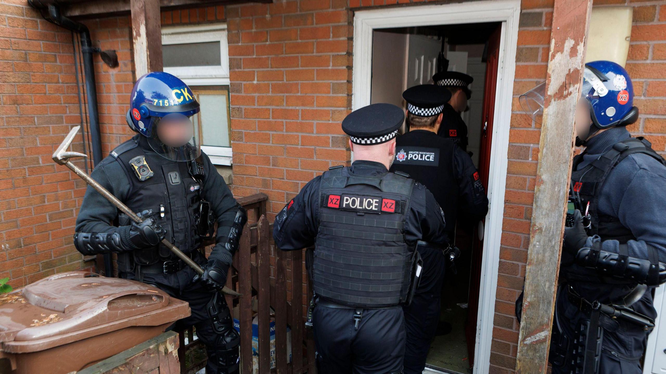 Officers in helmets holding crow bars stand either side of a door of a red brick home as three police officers walk inside.