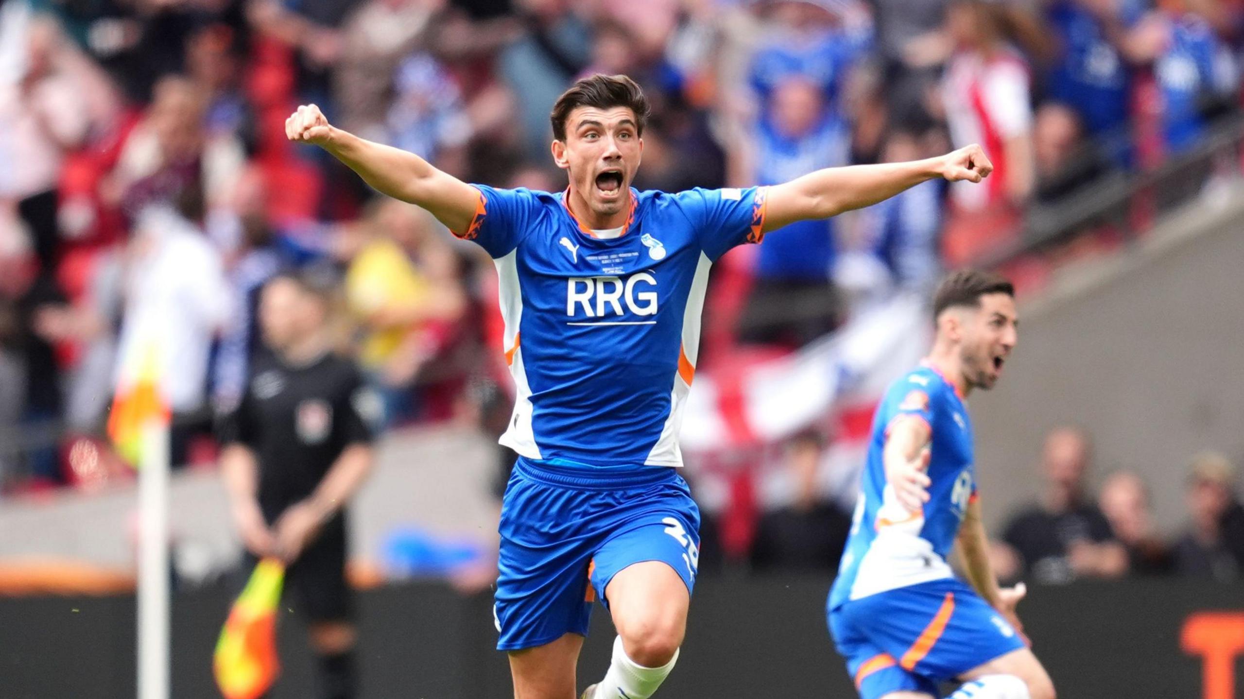 Kian Harratt is holding his arms in the air celebrating scoring a goal. He is wearing the Oldham Athletics kit.