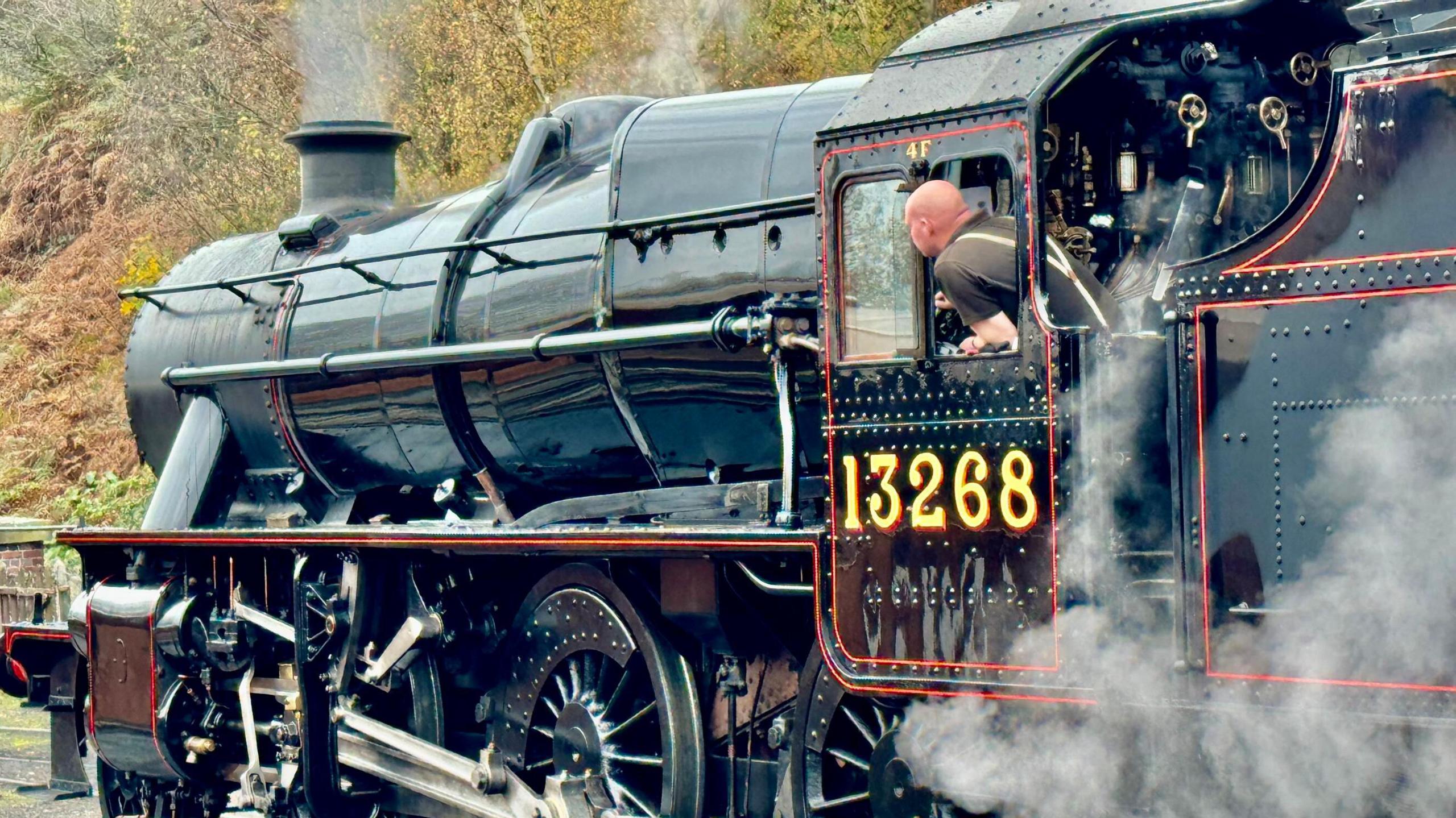 A close-up of a large black steam train on railway tracks. There is a man leaning out of its cab. There is steam coming from its chimney and near its wheels. There are yellow numbers on its side, reading "13268"