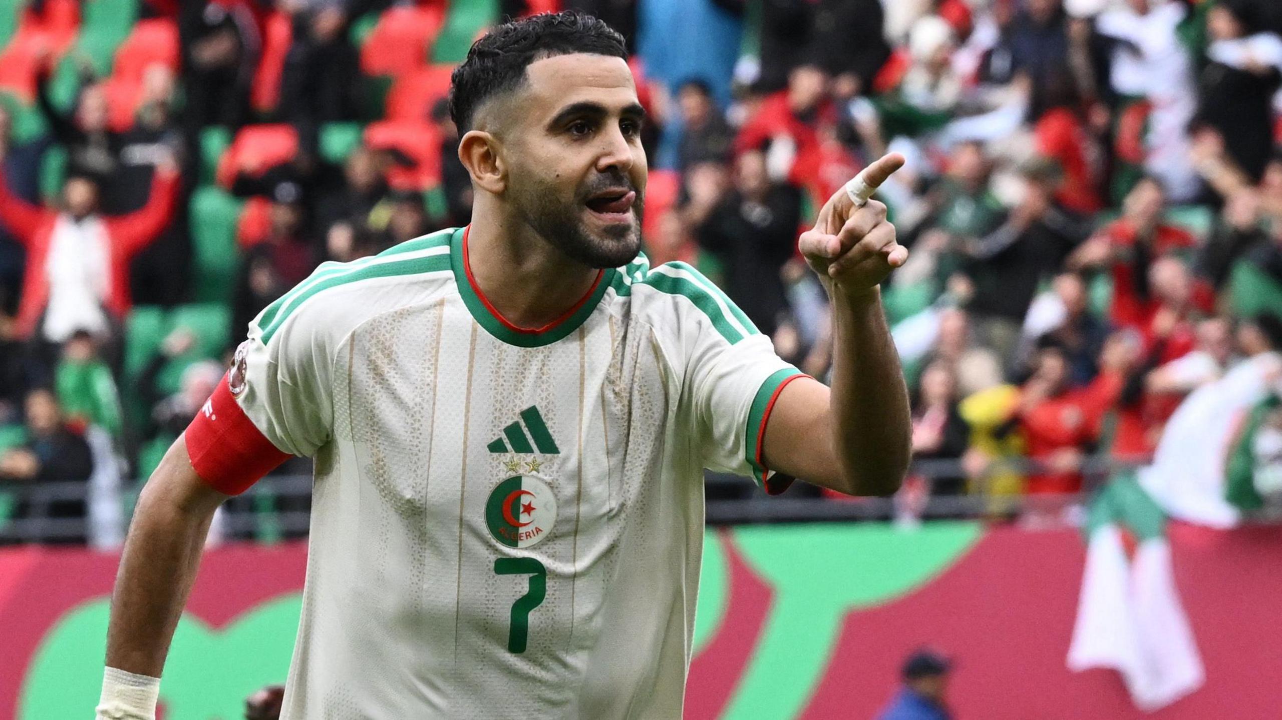 Riyad Mahrez, wearing a white Algeria shirt with the number 7 on the front, points with his left hand as he celebrates a goal for Algeria