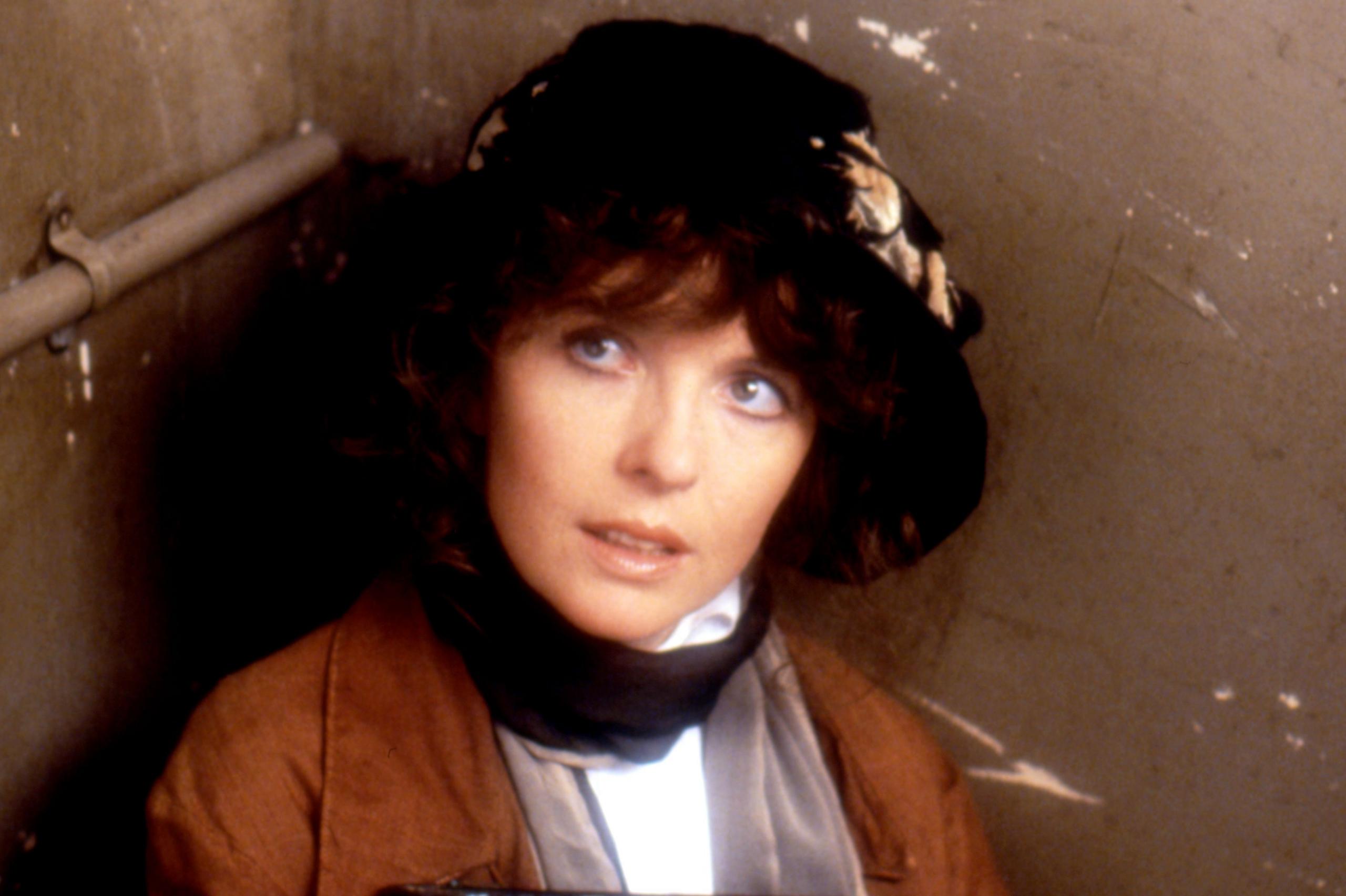 Diane Keaton e fotografuar me kostum gjatë xhirimeve të filmit të vitit 1980, Reds. Ajo është ulur në një hapësirë ​​të vogël të mbyllur me mure ngjyrë bezhë dhe një tub metalik të dukshëm që kalon përgjatë anës së majtë. Ajo ka veshur një pallto kafe mbi një veshje të bardhë dhe ka një kapelë të errët të zbukuruar me një shall ose fjongo me motive të çelëta. Ndriçimi është i zbehtë, duke krijuar një atmosferë të errët dhe të mbyllur.
