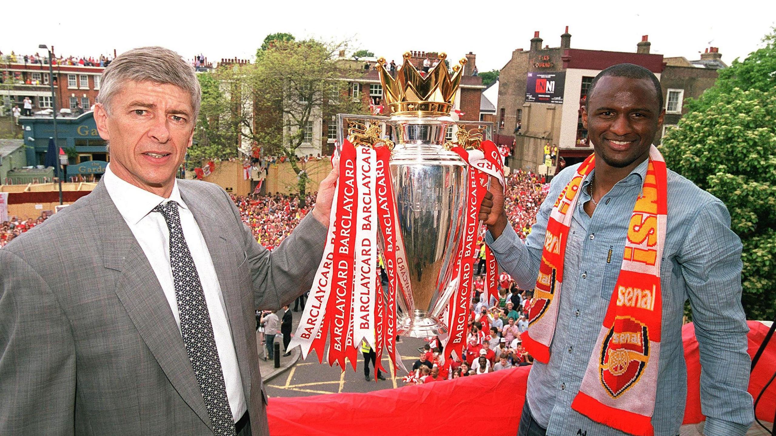 Arsenal manager Arsene Wenger and captain Patrick Vieira hold the Premier League trophy from a raised position as fans watch on from street level below.