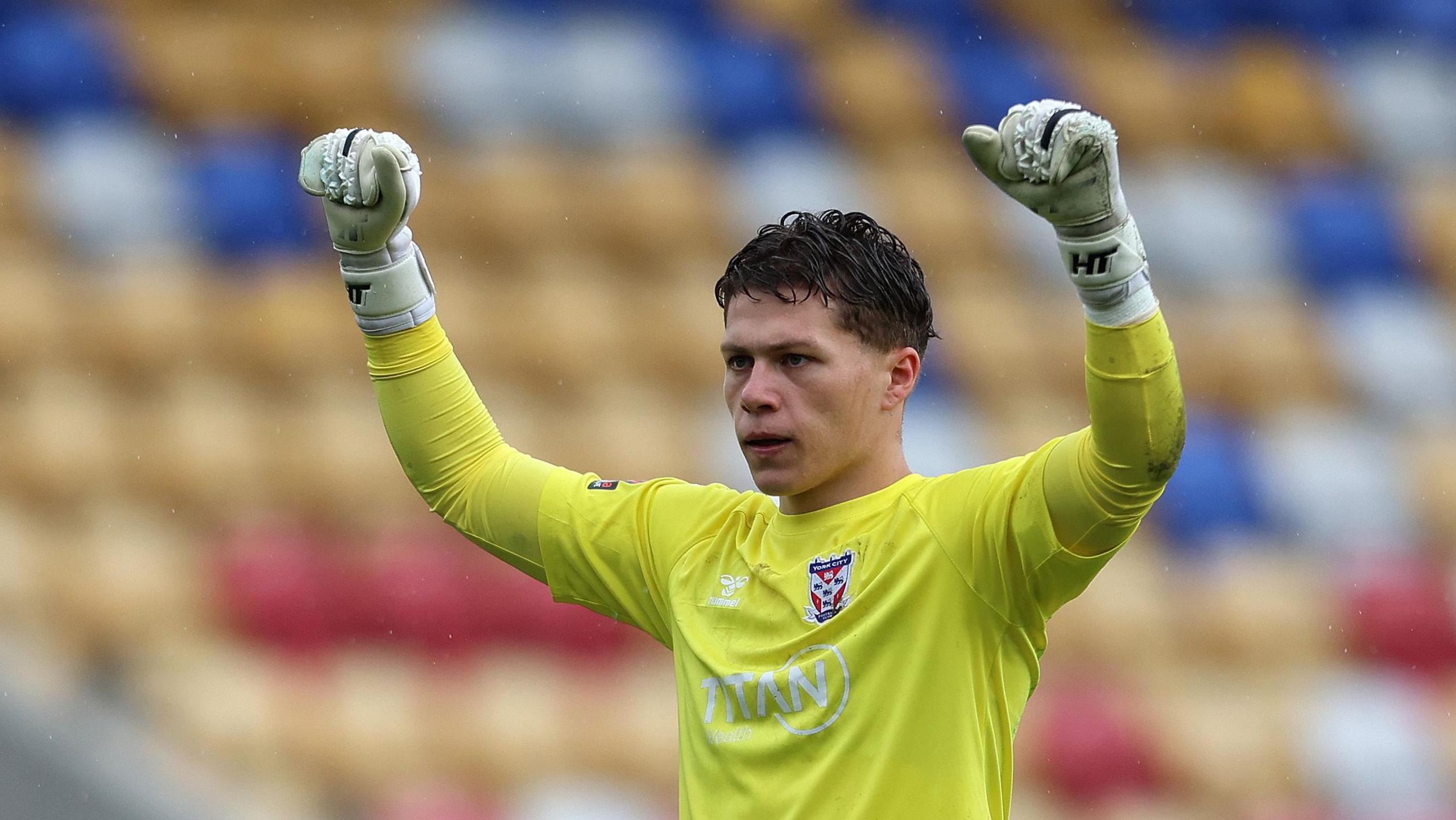 Harrison Male, wearing a yellow goalkeeper's jersey and white goalkeeper gloves, raises his hands in the air while playing for York City