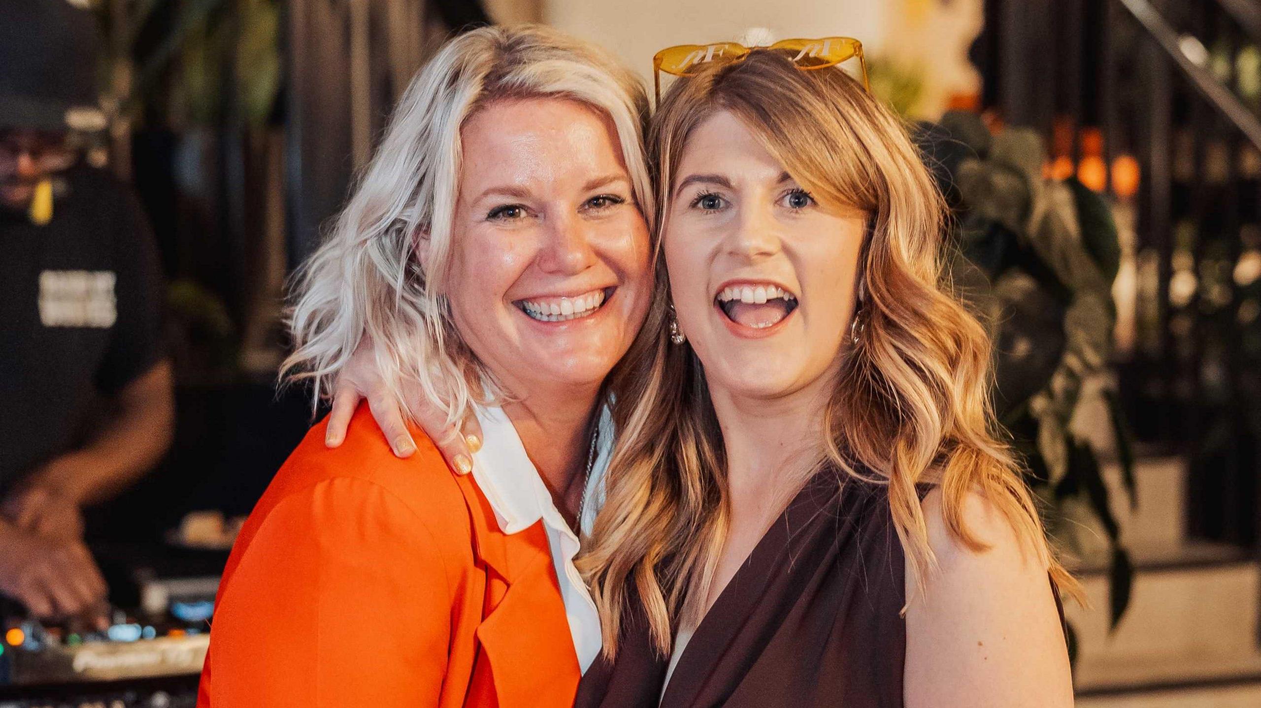 Two women stand cheek to cheek as they smile at the camera. One is younger and has blonde hair and a pair of orange sunglasses on the top of her head. She is wearing a plum-coloured dress. The other woman has fair hair and is wearing an orange blazer and white shirt.