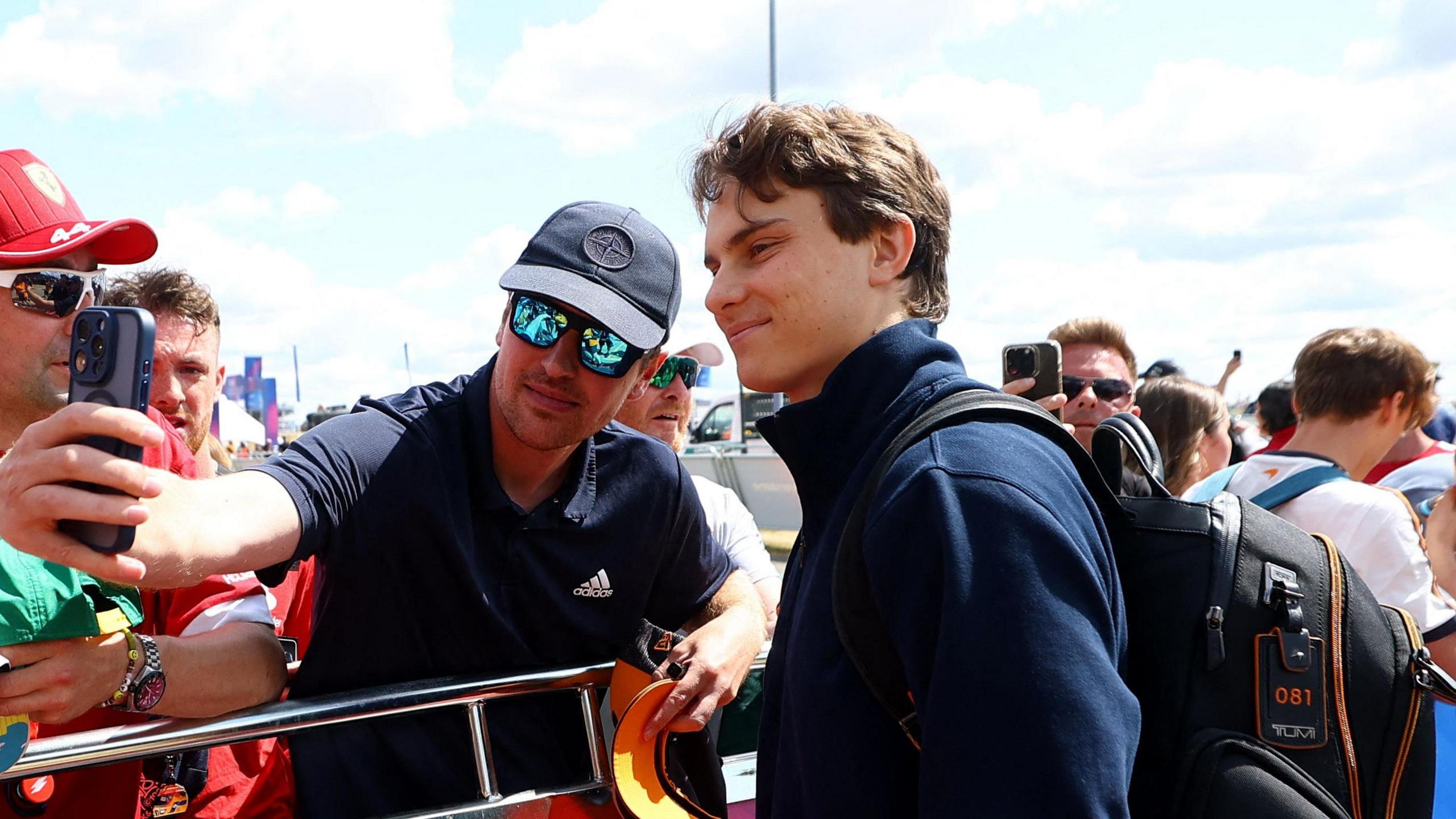 Oscar Piastri is just off centre to the right, looking to the left of the image. He is standing next to someone, who is on his left, taking a selfie. They are both wearing a navy T-shirt.