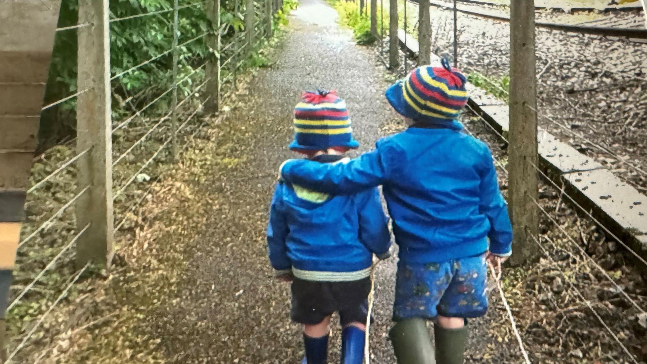 Two little boys in blue jackets with knitted hats with red, yellow and white stripes, wearing wellies walking toward the railway station 