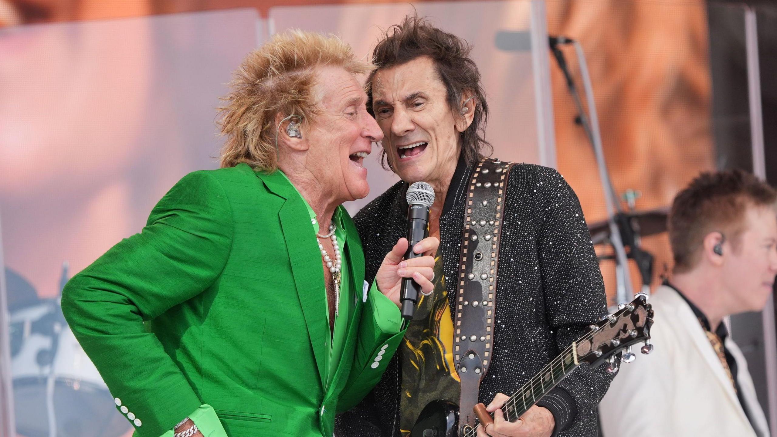 Two men are on stage singing into the same microphone. One of them is wearing a green suit, the other a black jacket.