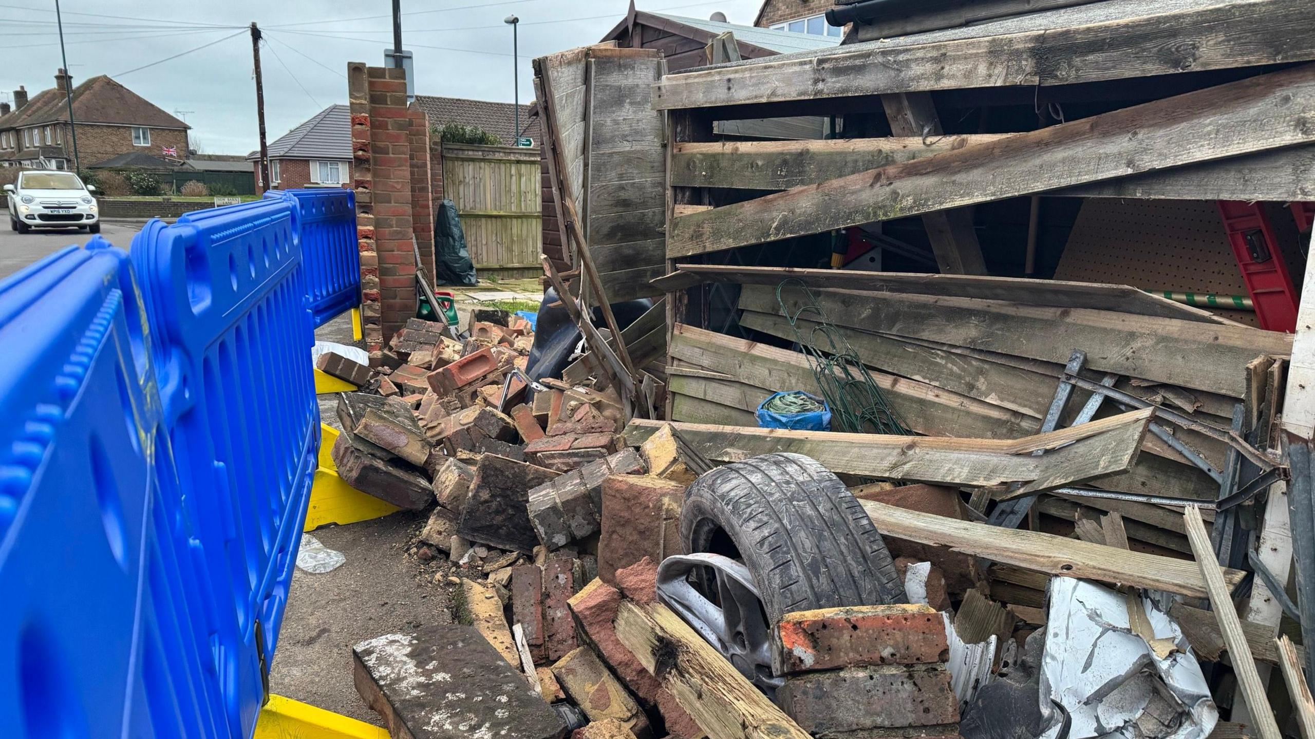 Bricks still scattered on the floor following the incident on 30 January. One of the car's wheels and part of its bumper are still lodged among the debris. A blue plastic fence is set up on the left to cordon off the debris from the road and part of a wooden shed is also damaged. 