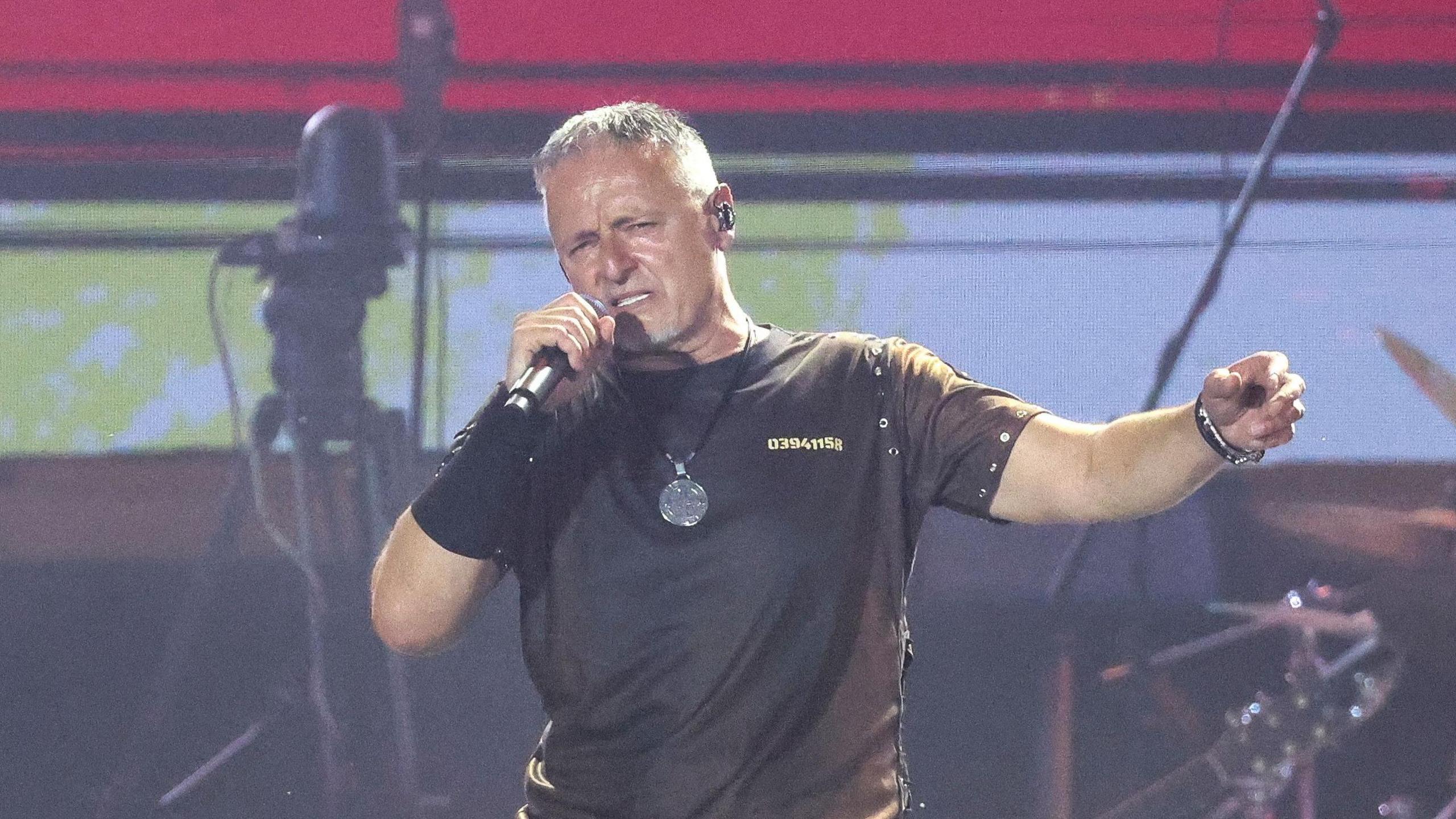 Marko Perkovic on stage at this month's concert. He is a 58-year-old man with grey hair wearing a black t-shirt and a necklace with a large silver medallion.