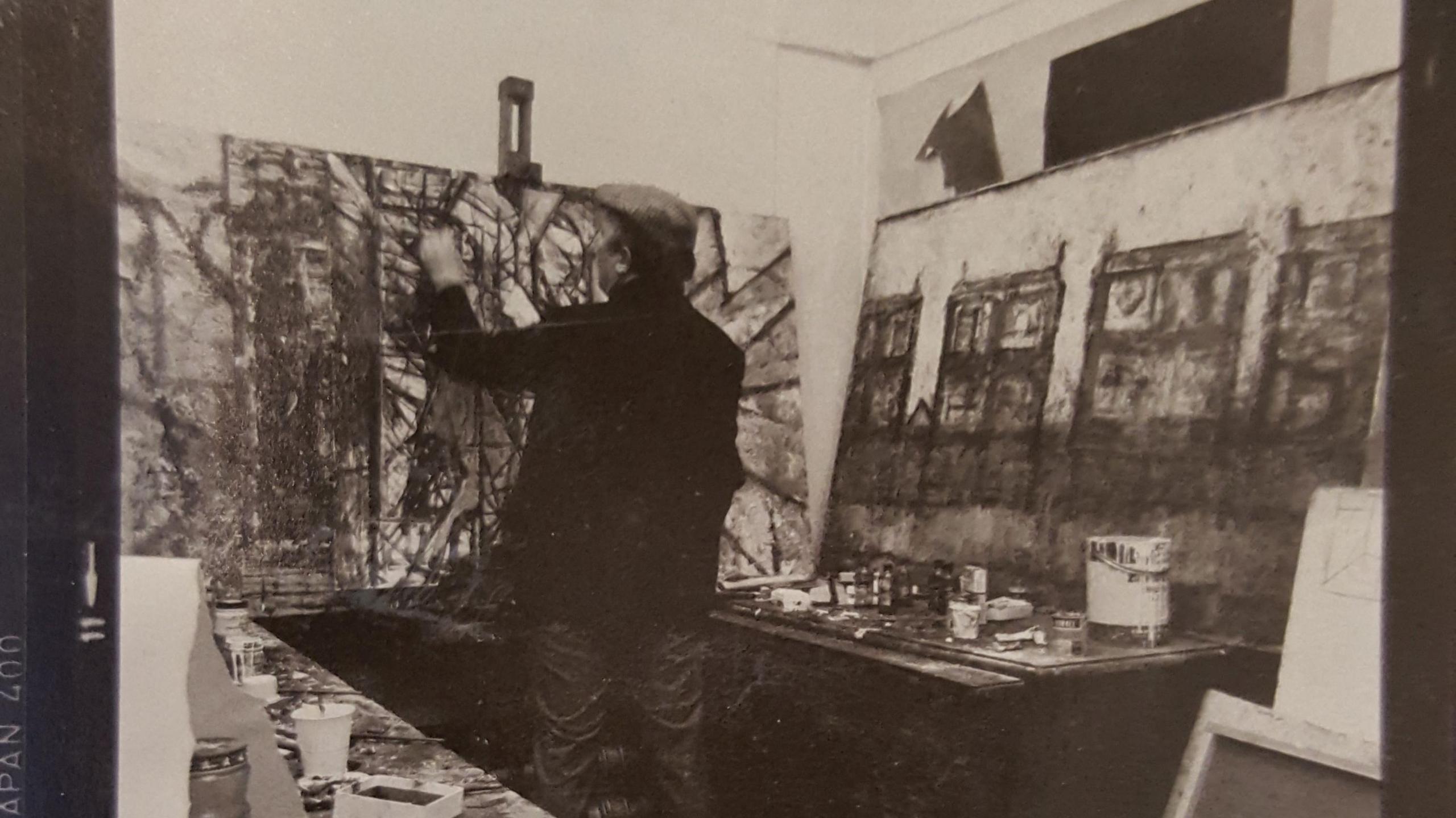 A black and white image of a man in dark trousers and a dark coat wearing a hat. He has his back to the camera and is inside an art studio. He is  drawing on a large canvas which is standing up on a workbench, which has black geometric shapes on it.