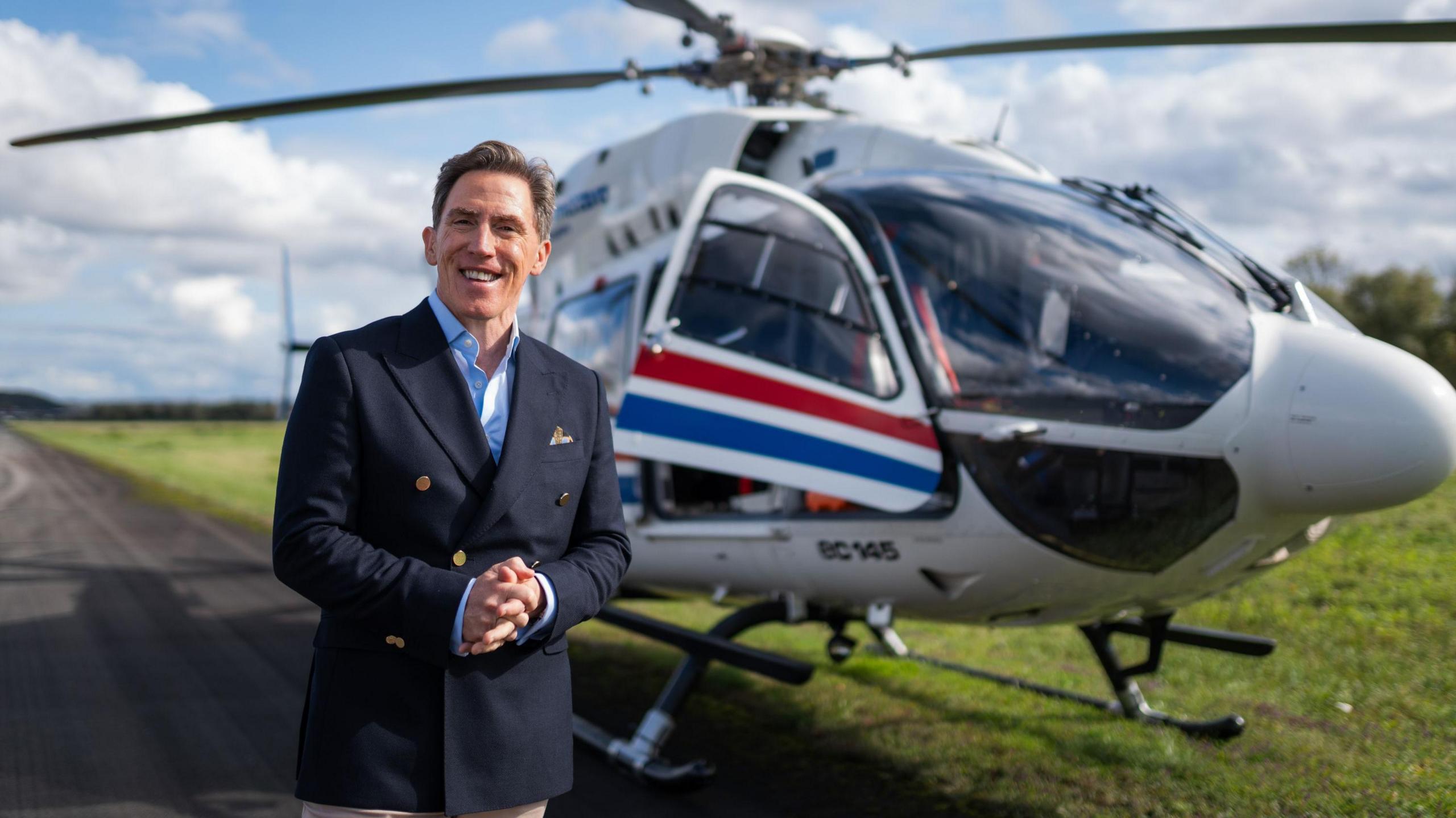 Rob Brydon wearing a blue suit jacket with a blue shirt underneath. He is stood in front of a white helicopter with red and blue stripes on the doors.