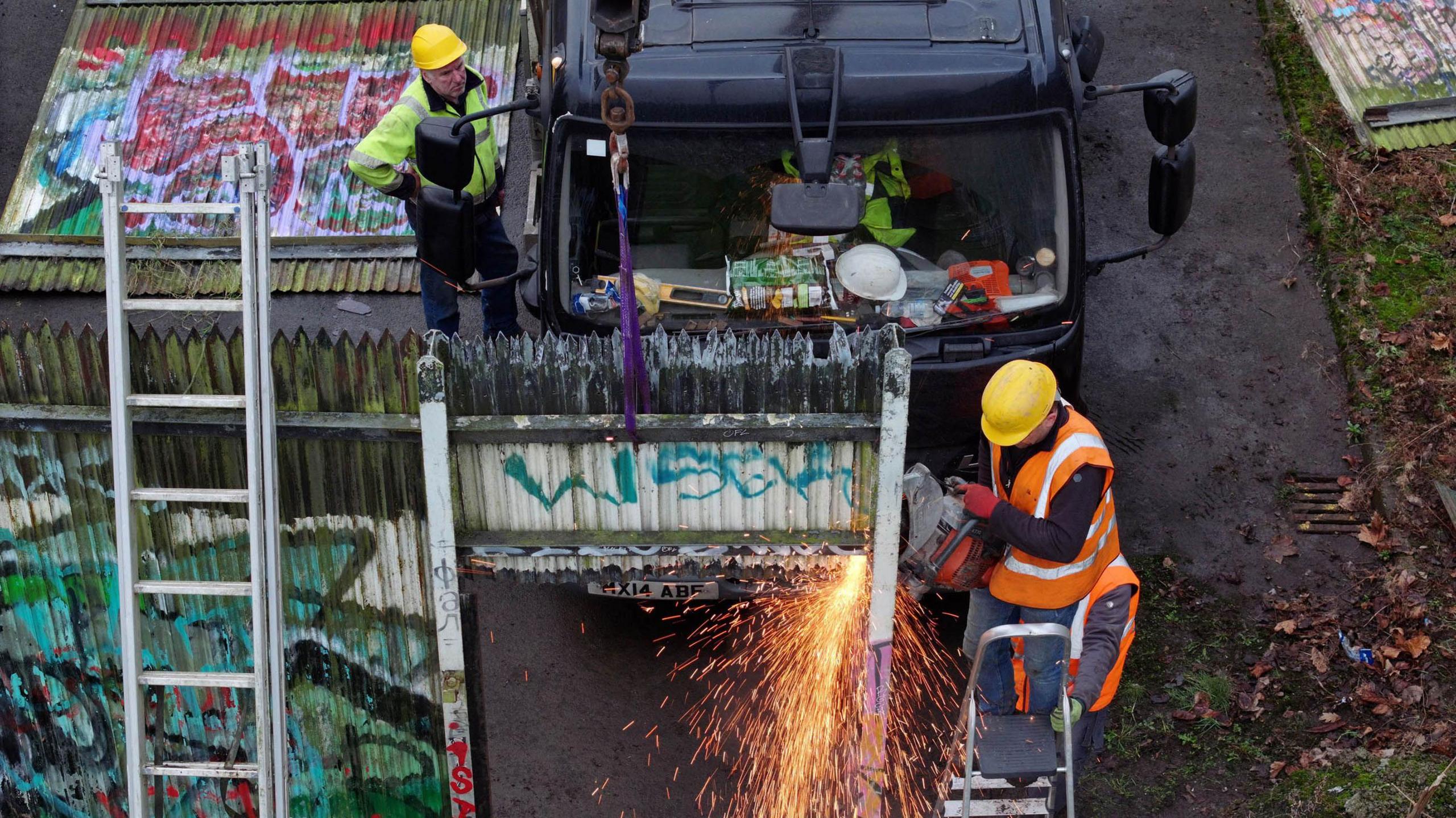 Two men in Hi-Viz jackets dismantle a steel wall that has been covered in multi-coloured graffiti. The wall sits at the end of a street with a black lorry parked behind them. One of the men is on-top of a ladder with a steel cutter in hand. Sparks can be seen flying of the cutter. 