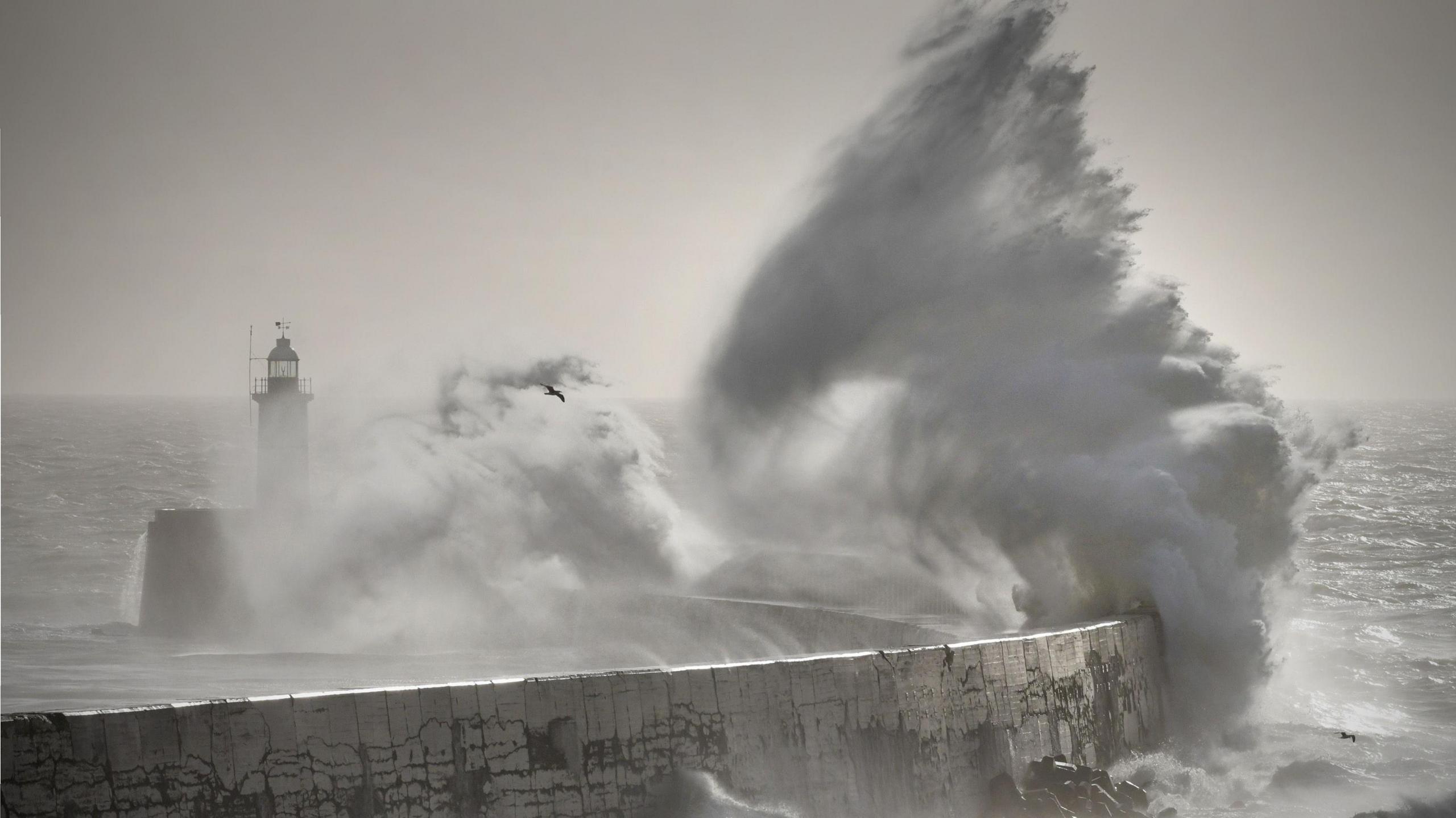A huge wave crashes over a breakwater and lighthouse, with grey skies overhead and stormy seas in the background