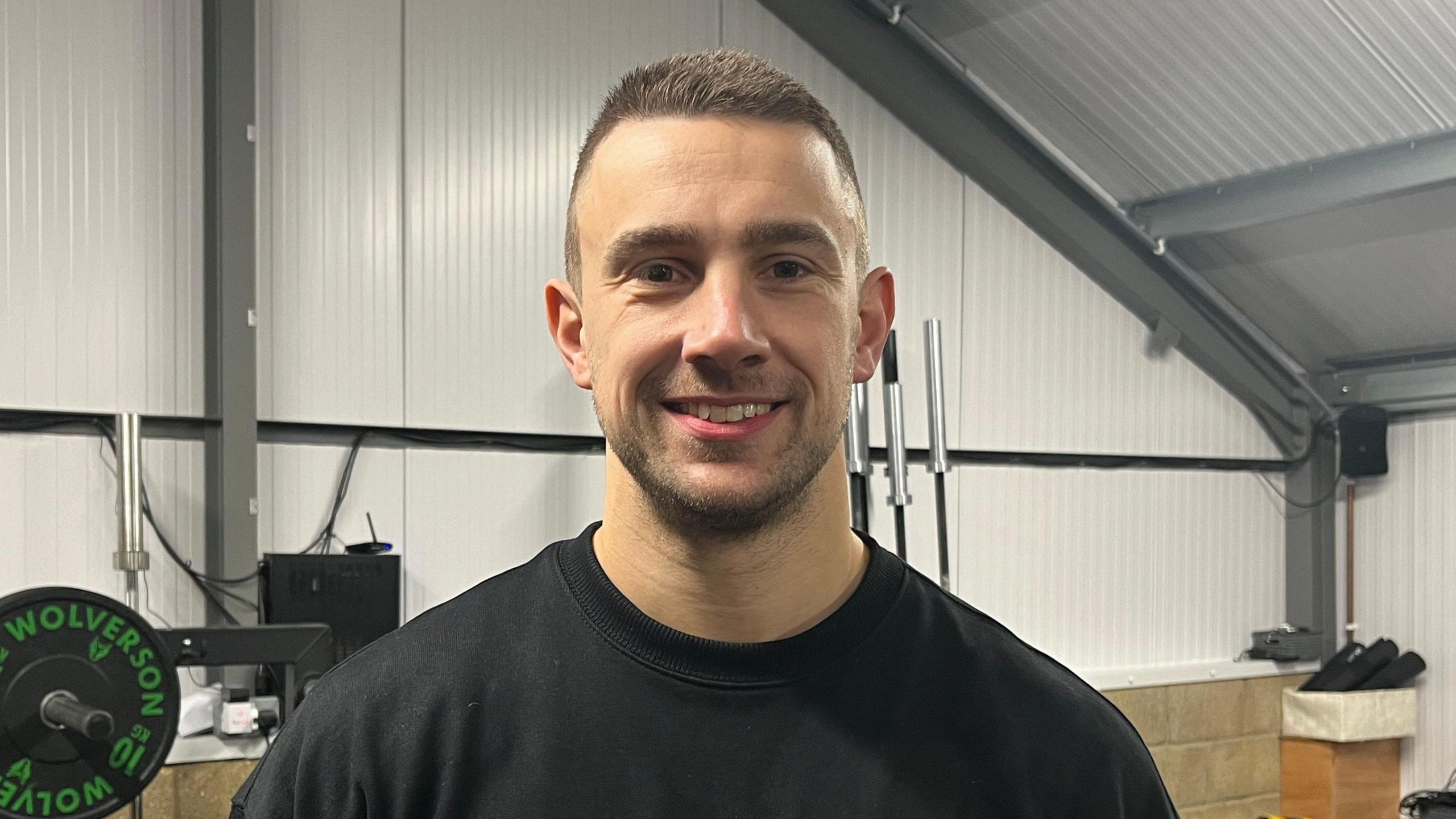 Mike is smiling at the camera and wears black gym pants, a black sweatshirt with Overload  in the centre and has short dark hair and facial hair. He has brown eyes and is stood in front of weights and gym equipment. 