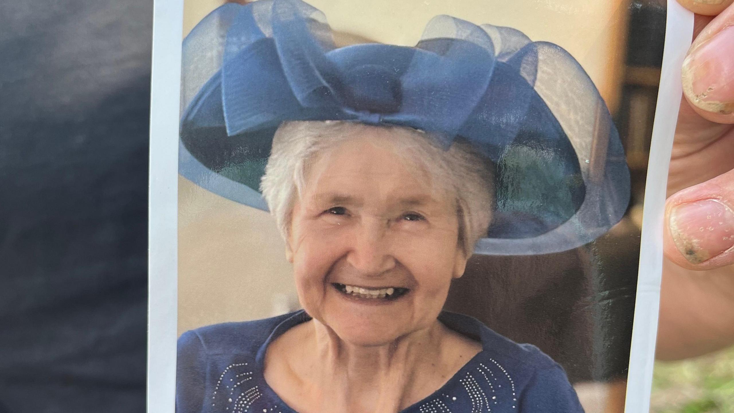 Carol is wearing a blue dress and hat. She is smiling, looking into the camera.