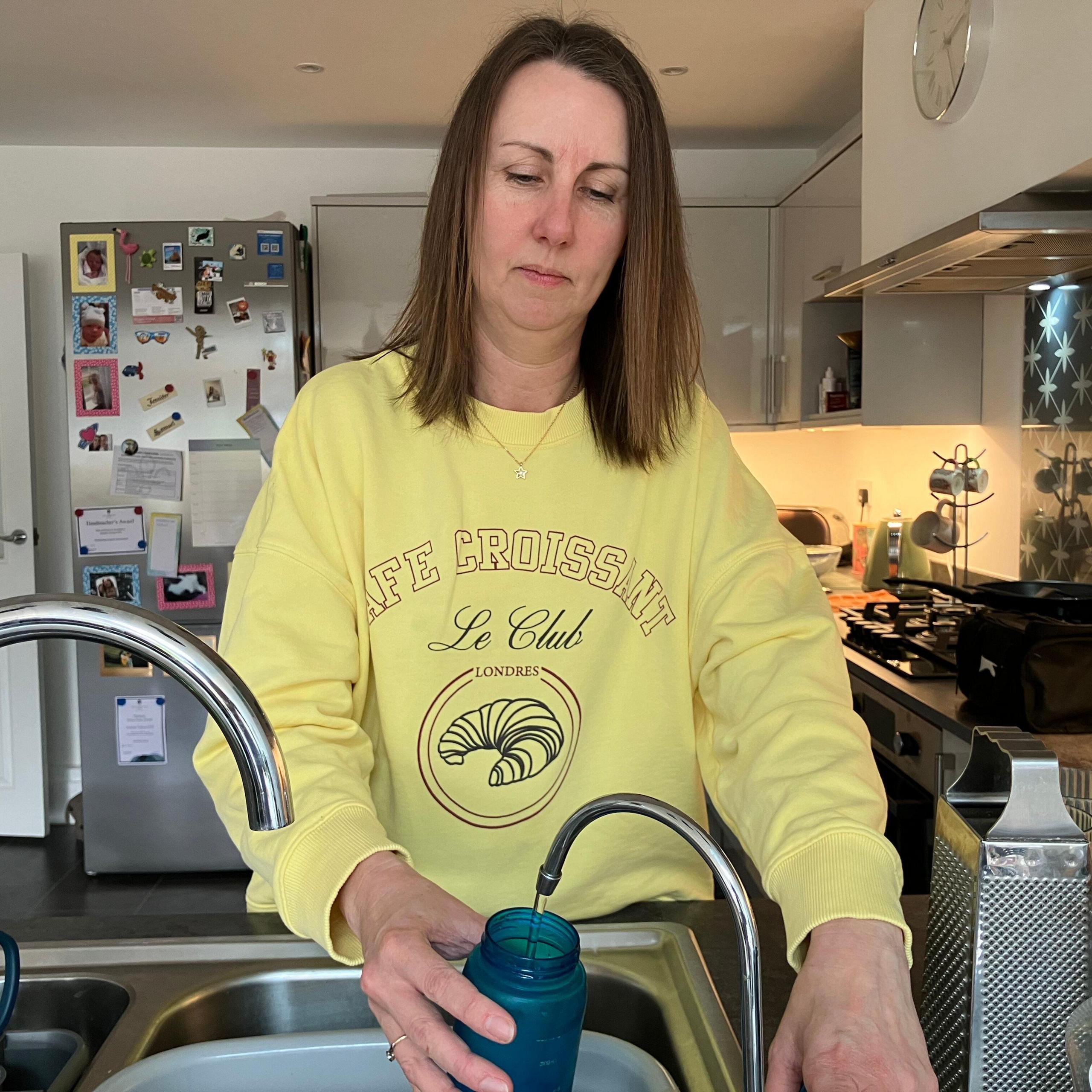 A woman with medium length brown hair wearing a bright yellow jumper pouring water from the sink into a blue water bottle.