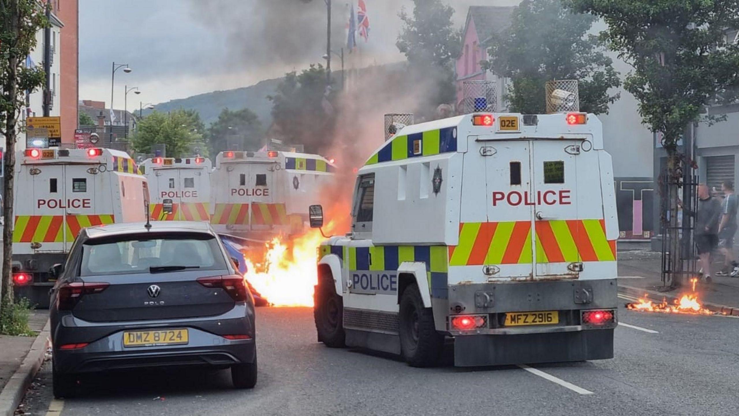 A police landrover seen from the back with a fire in the middle of the road ahead of it. Three other land rovers in the background