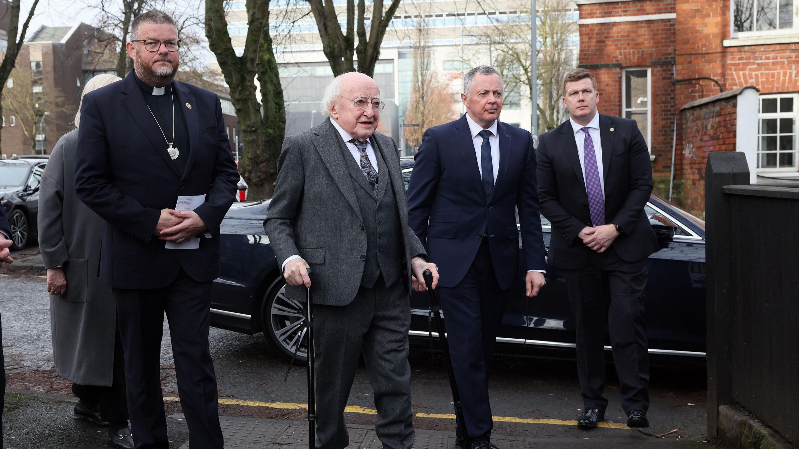 Left to right:
A minister/priest wearing the collar and black suit clutching an order of service. He has glasses and short hair. 
Michael D Higgins wearing grey tails, a white shirt and grey tie. He has white hair and has a walking stick in his right hand.
Two unknown men in black suits, one with a blue tie and one purple. The left one has grey hair and the right one has ginger hair.