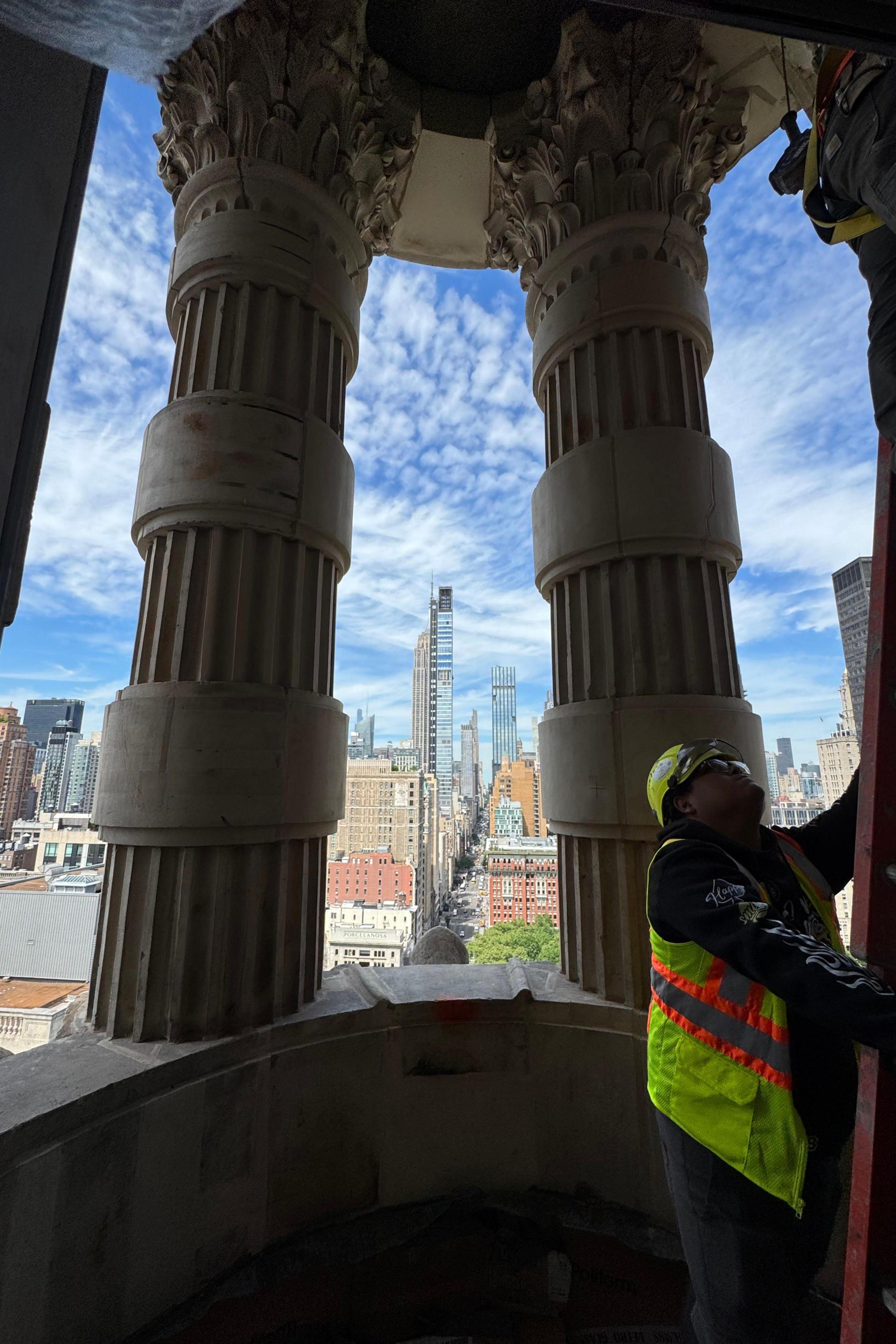 Image of construction worker in yellow vest with a ladder on a balcony with large columns and a view of the Manhattan skyline