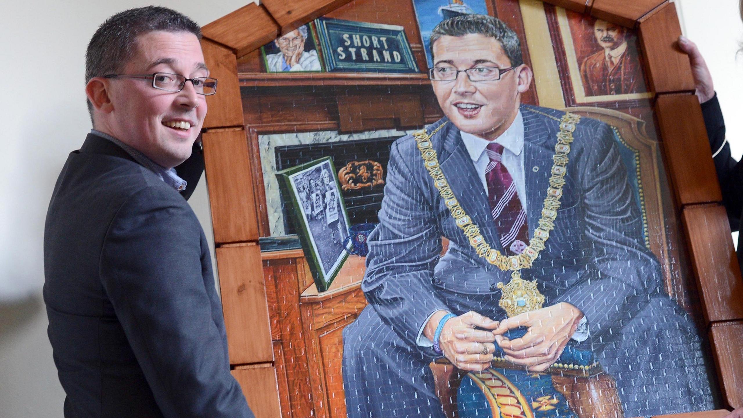 Niall Ó Donnghaile carrying his portrait. He is wearing a navy suit, blue shirt and red jumper. 