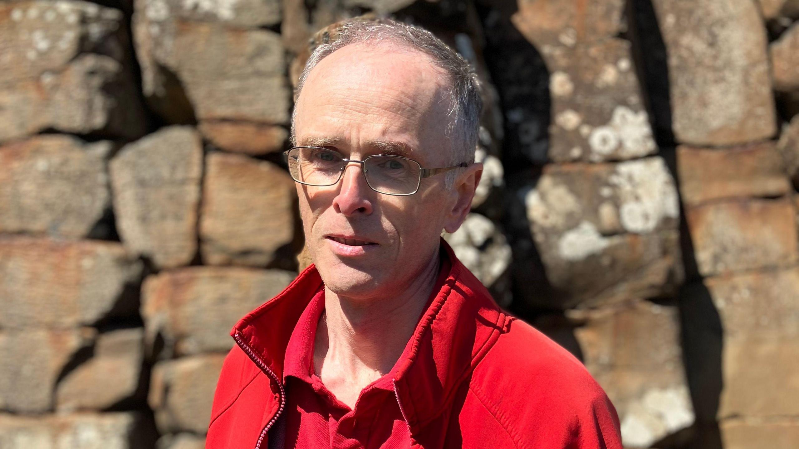 Dr Henry is looking at the camera and is wearing thin framed glasses. He has thin grey hair and is wearing a bright red fleece and polo top.