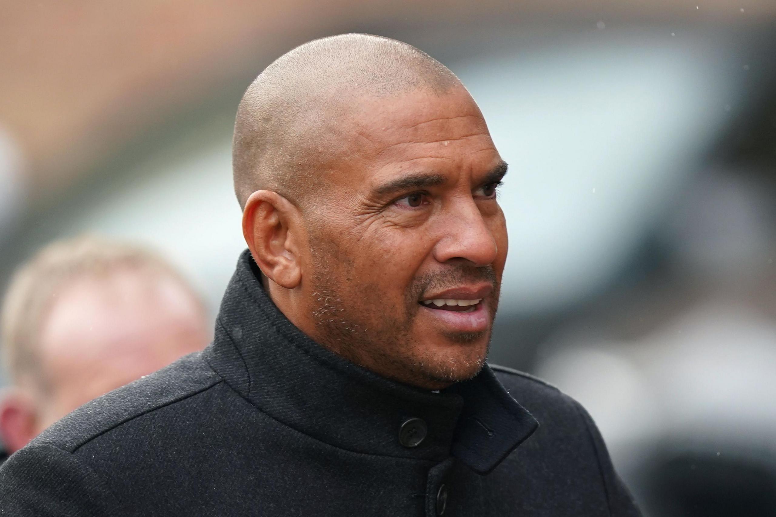 Stan Collymore wearing a black coat looks to the right of the image. He is standing outside.