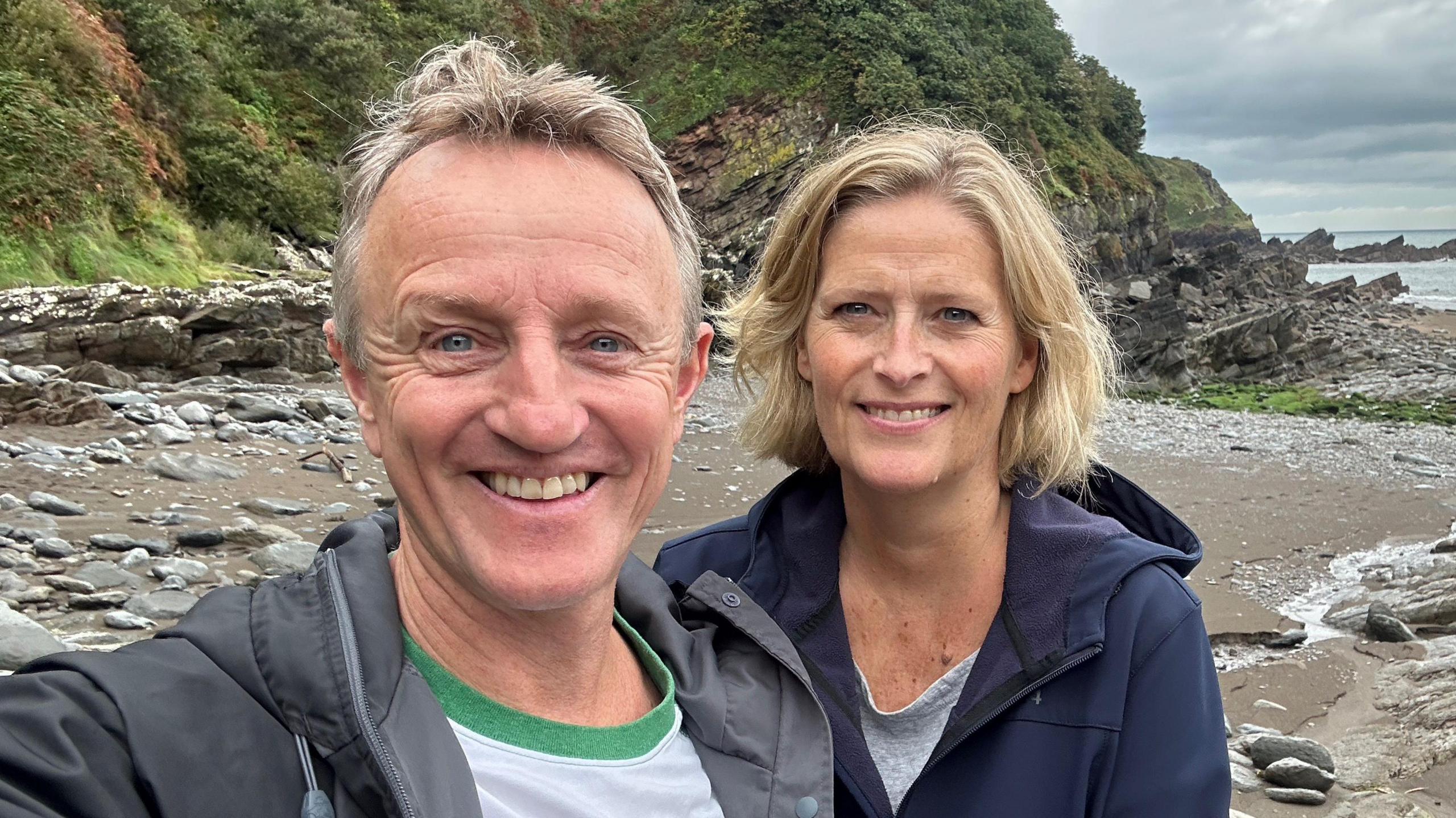 Peter Lloyd-Williams in a white shirt and black coat standing on a beach next to Fiona, who has shoulder-length blonde hair and a navy coat on.