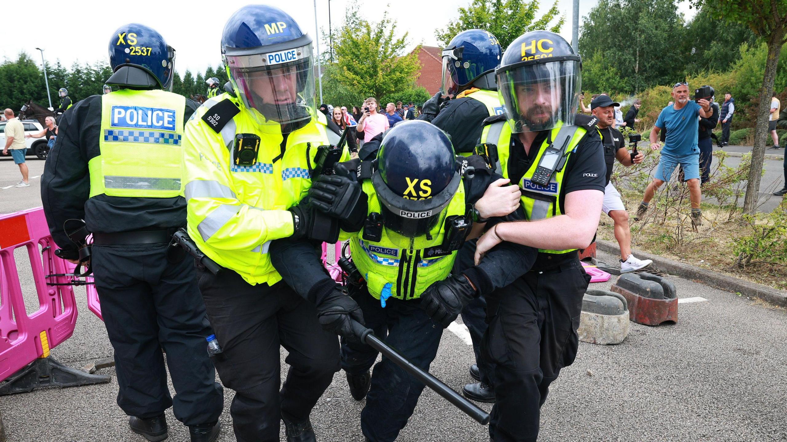 Police officers wearing high-vis jackets with helmets and face shields. Two police officers flank a third who is being held up. 