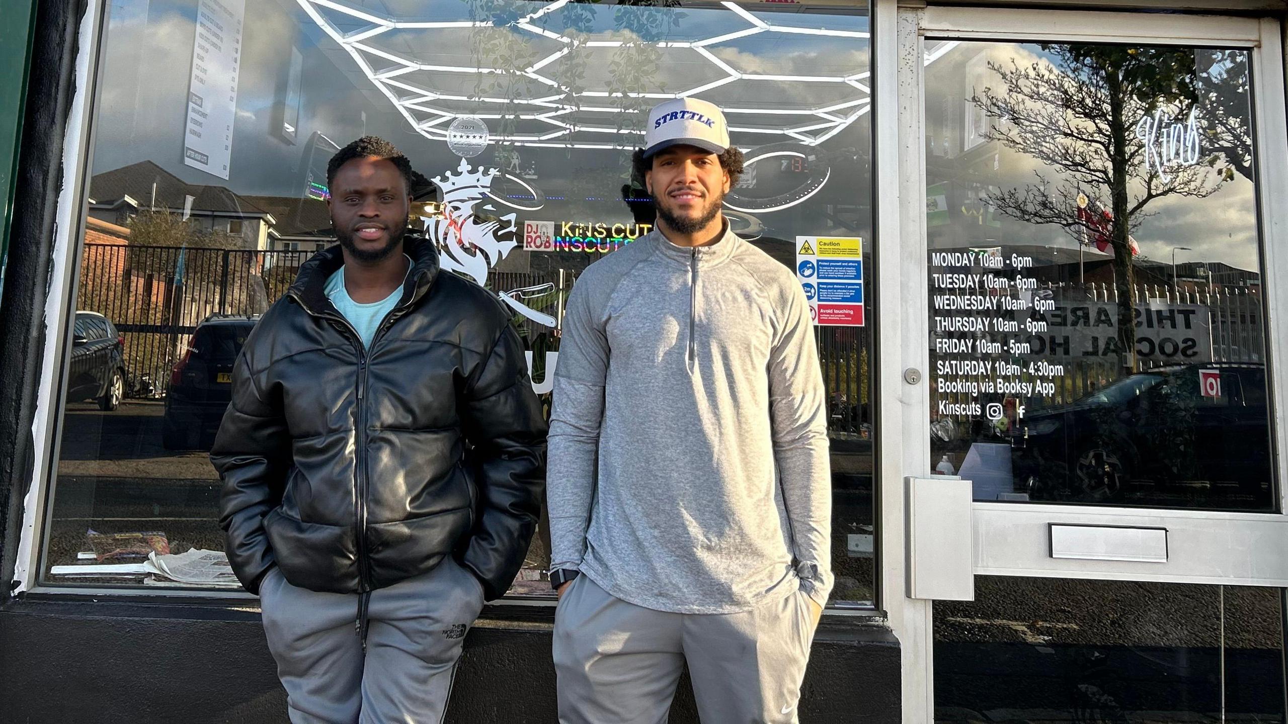Two men stand in front of a barber shop window with their hands in their pockets. It is a pale yellow building and the barber shop is painted green around the door and window. The man on the left is wearing a black coat and grey trousers. The man on the right is wearing a hat, grey top and trousers.