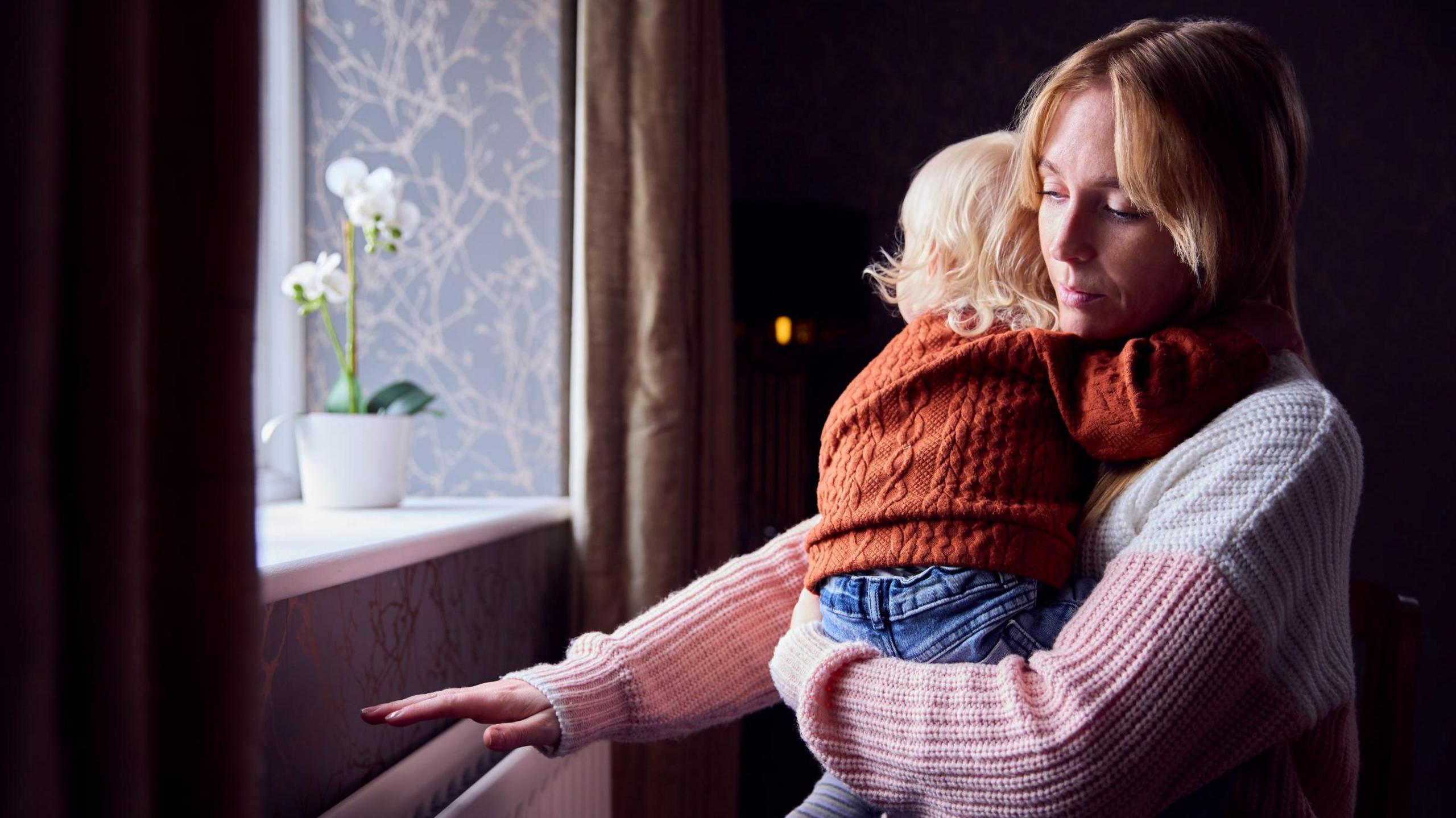 A woman is trying to stay warn while holding her young child - she has one hand over a heater at the window while her other arm is holding the child up. She has fair hair and a white and pink jumper on. The child has their back to camera but are looking out a window. They have an orange jumper on, and blonde hair.