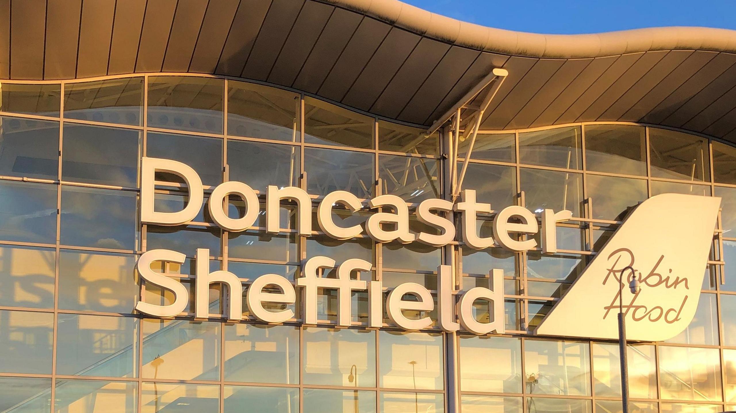 A large wall covered with glass windows has large white letters saying Doncaster Sheffield. There is a large white plaque shaped like an aeroplane tail which says Robin Hood