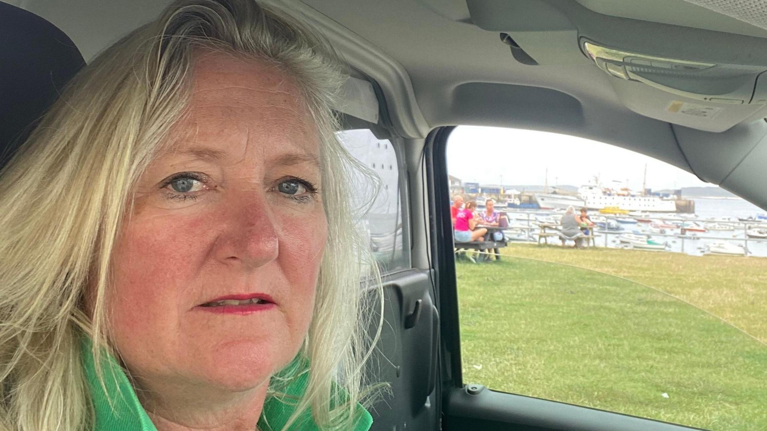 Sam sitting in a car with grass and a harbour in the background. She has long blonde hair, red lipstick and is wearing a green top.