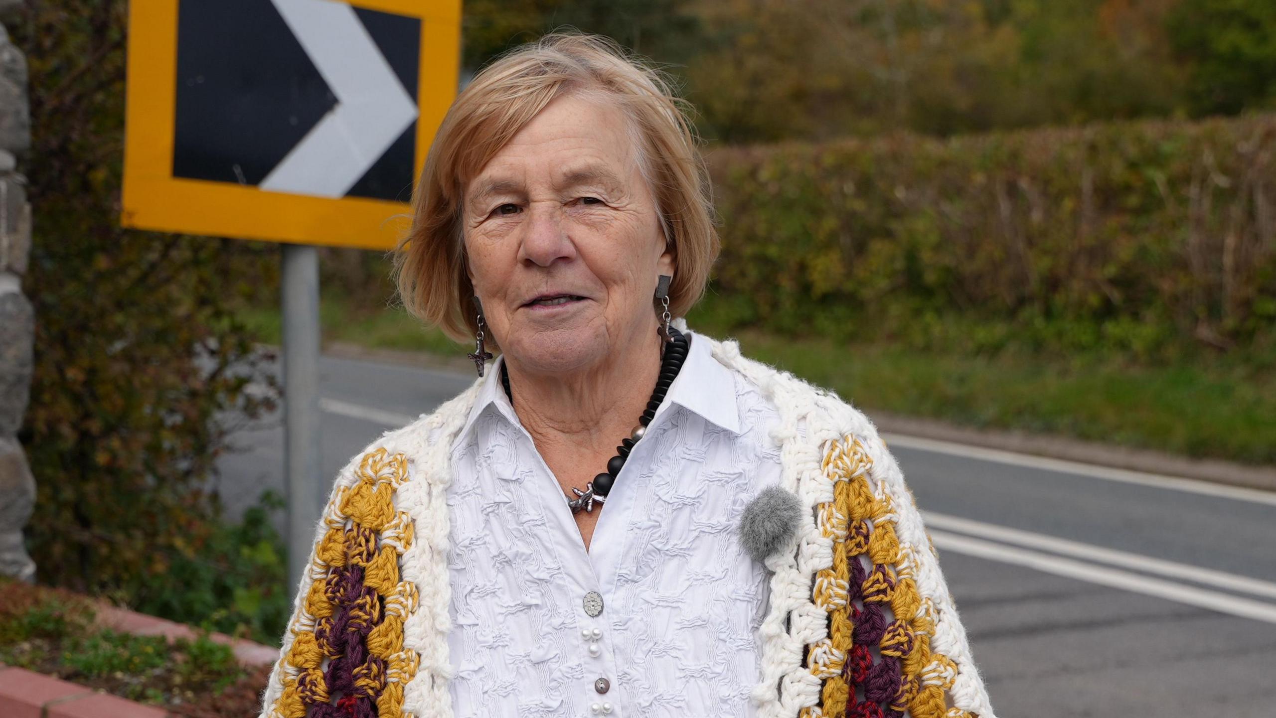 A woman with short reddish hair, wearing a white top looks at the camera. She is stood outside and there is a road sign behind her. It is a head and chest shot. 