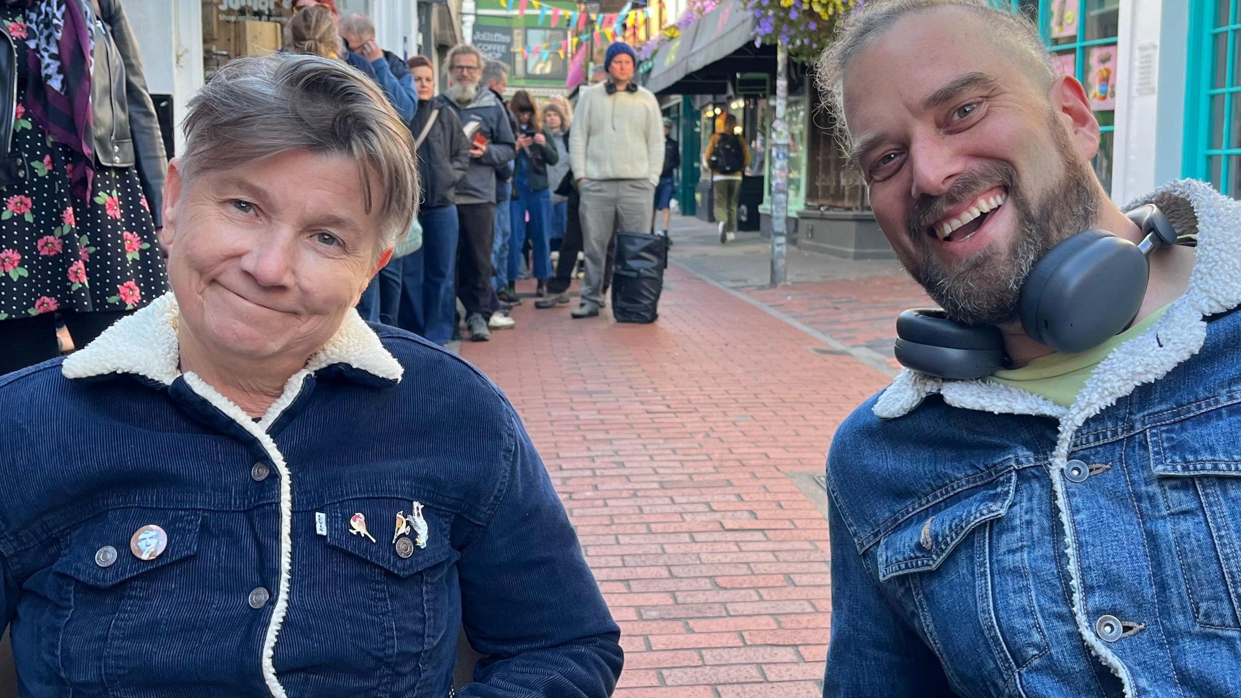 Zoe Cutting and Simon Smith queuing outside Resident Music in Kensington Gardens, Brighton and Hove. Both are wearing button up jackets; Zoe's is corduroy and Simon's is denim. They are both sitting in camp chairs.
